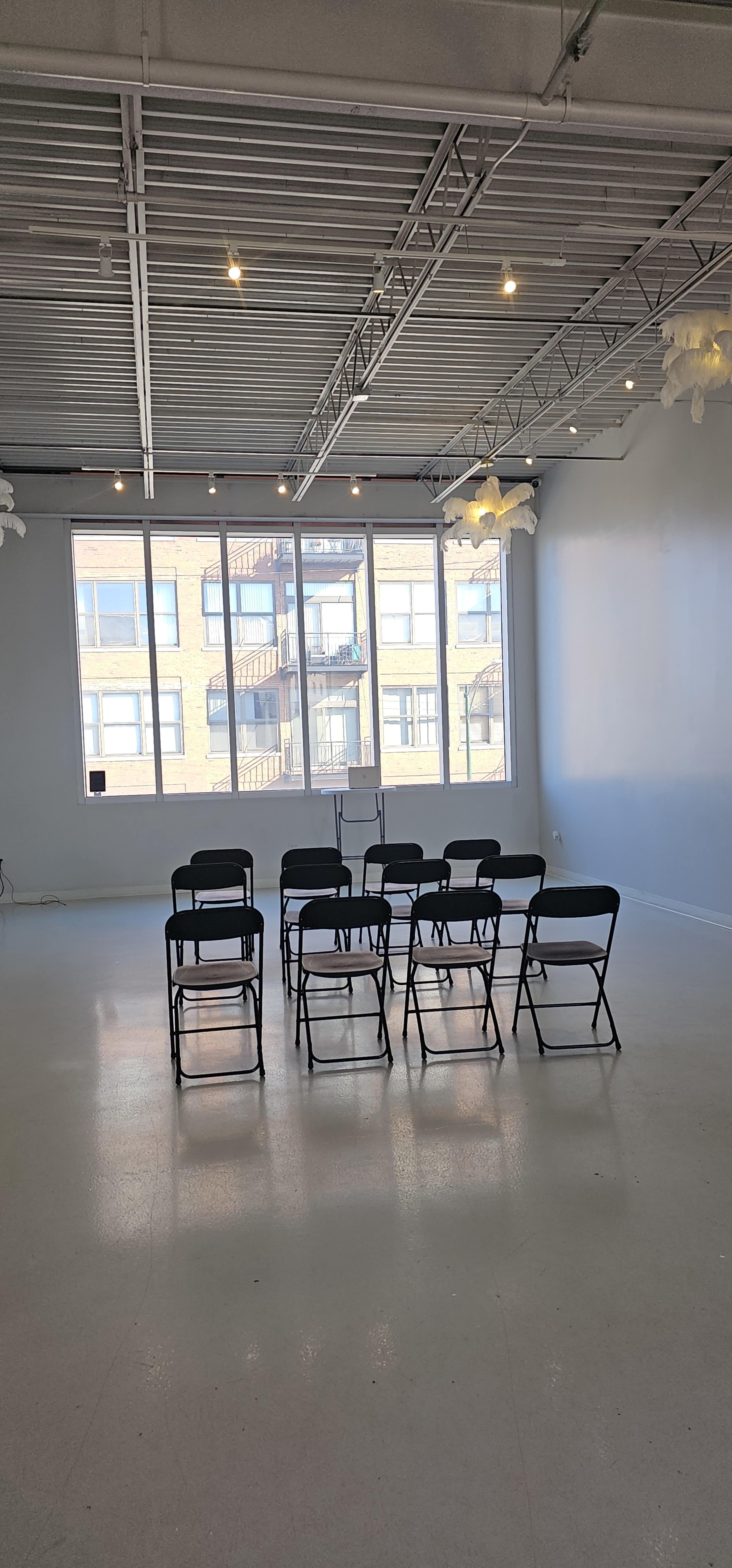 A row of black folding chairs is arranged facing a large window in a light-filled, empty room with white walls and a high ceiling.