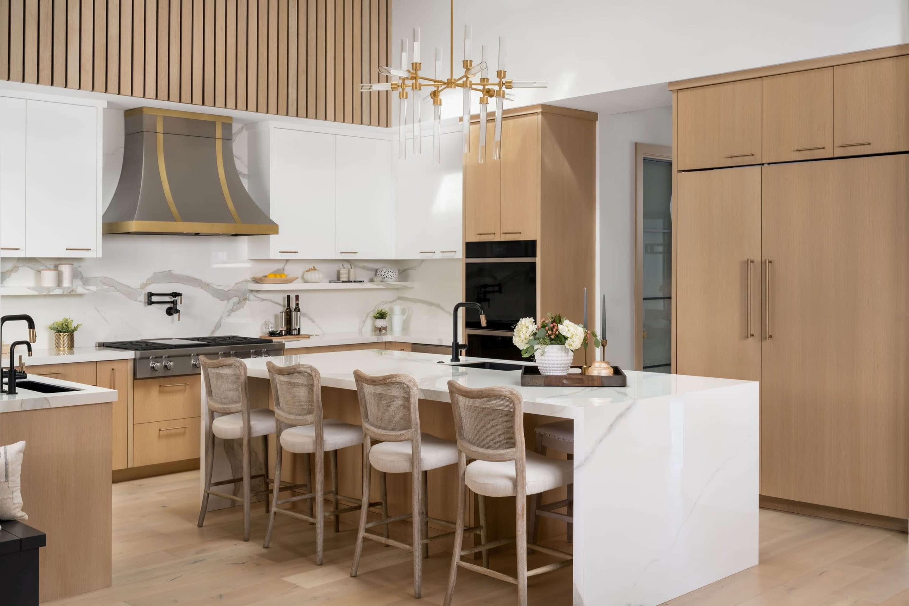 The image depicts a modern kitchen featuring a central island with bar seating, a gold-accented hood, and light wood cabinetry.