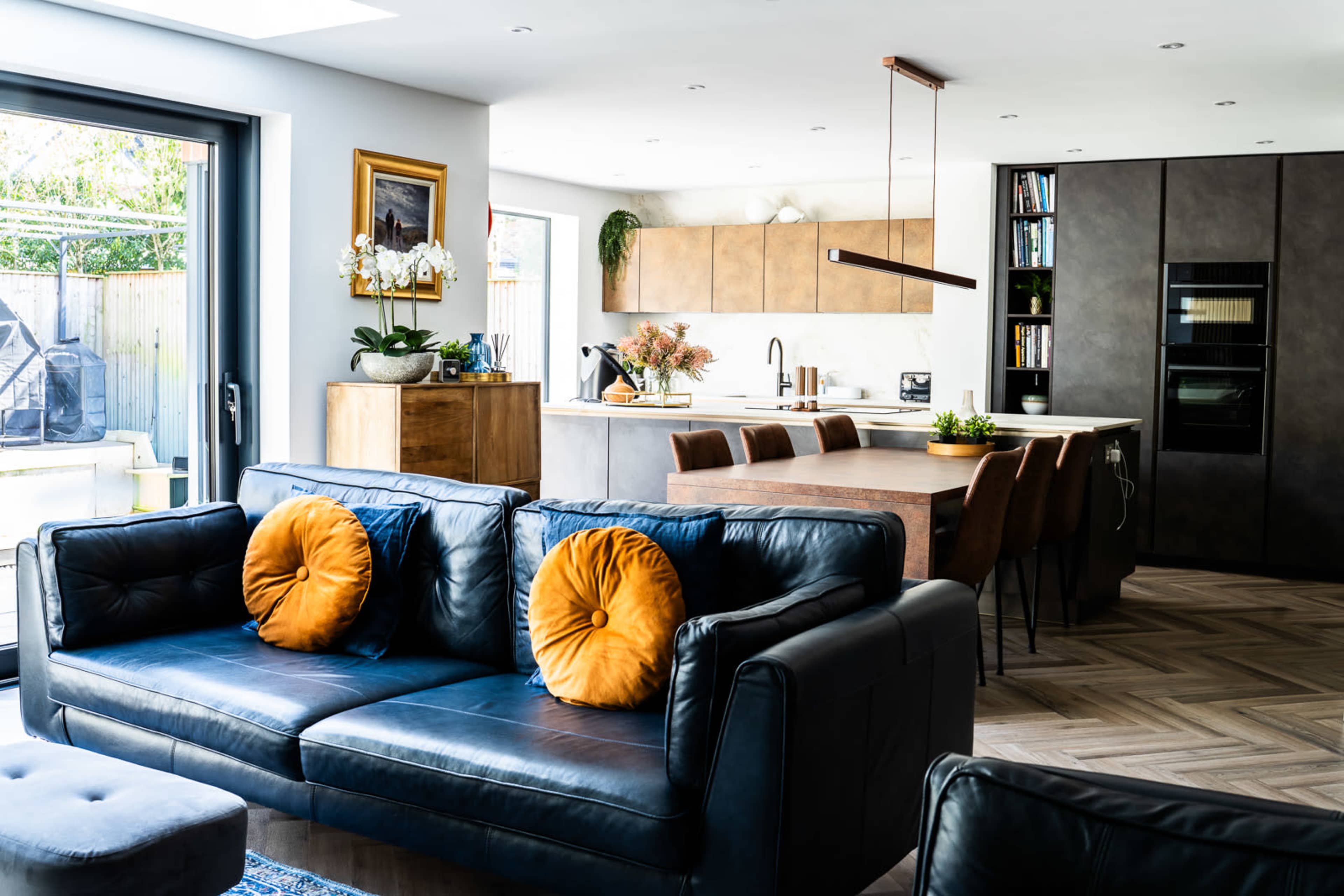 The image shows a modern living space featuring a black leather sofa with orange pillows, a wooden dining table, and a sleek kitchen area with a large window and contemporary decor.