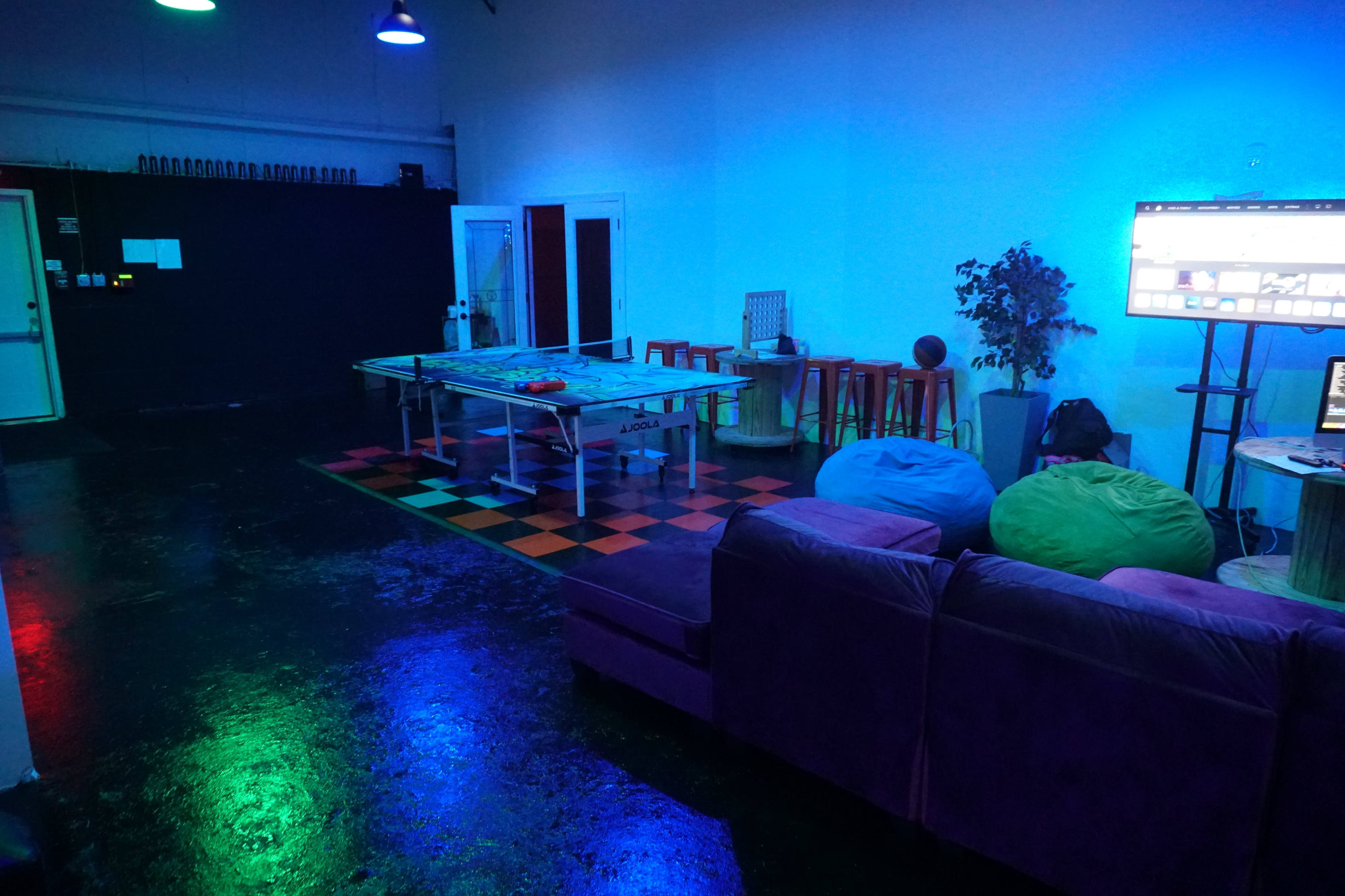 The room features a ping pong table, colorful bean bags, a couch, and a large screen set against a backdrop of blue lighting and black walls.