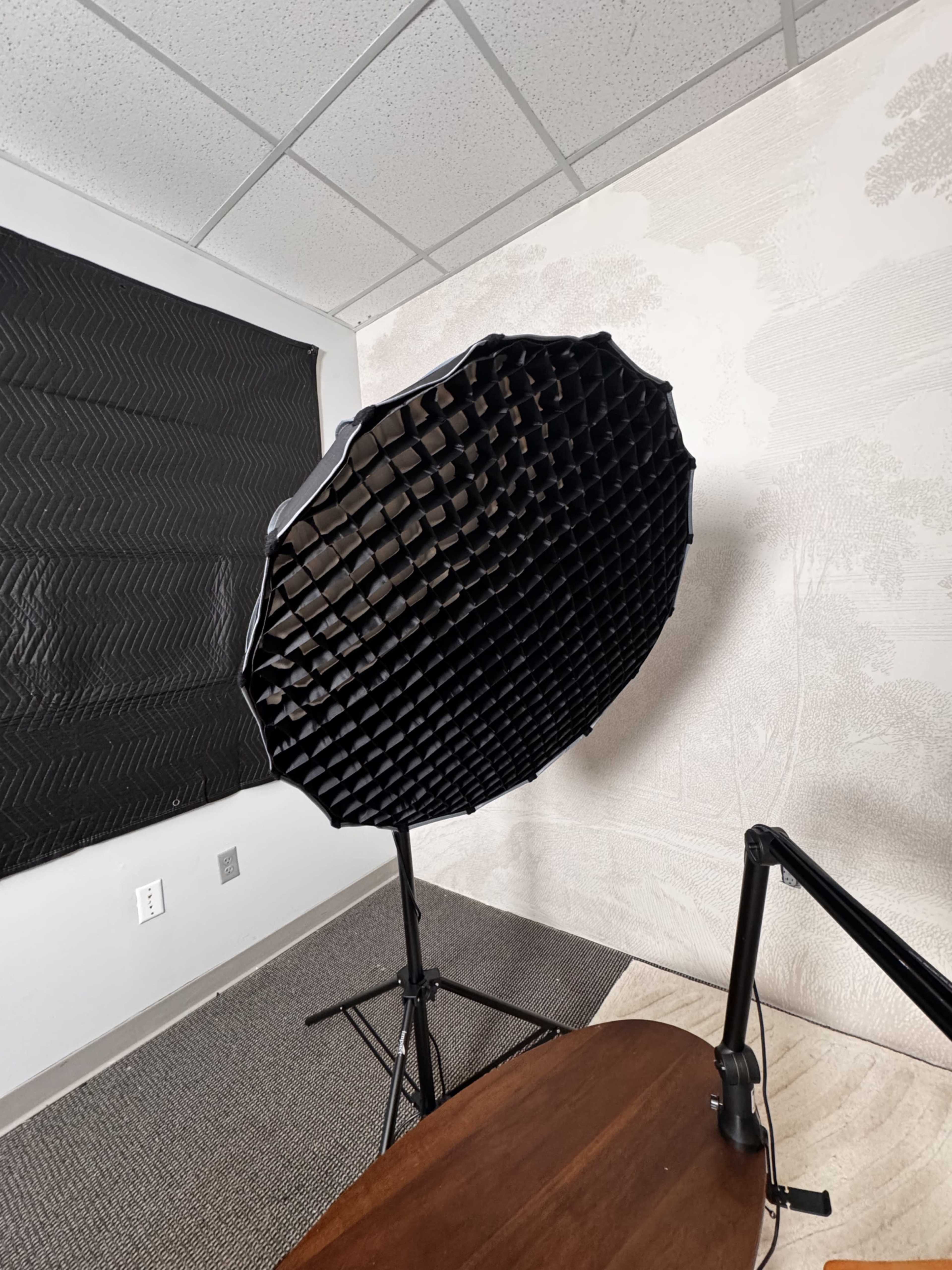 A large softbox with a grid attachment stands on a tripod in a well-lit room adorned with patterned wallpaper.