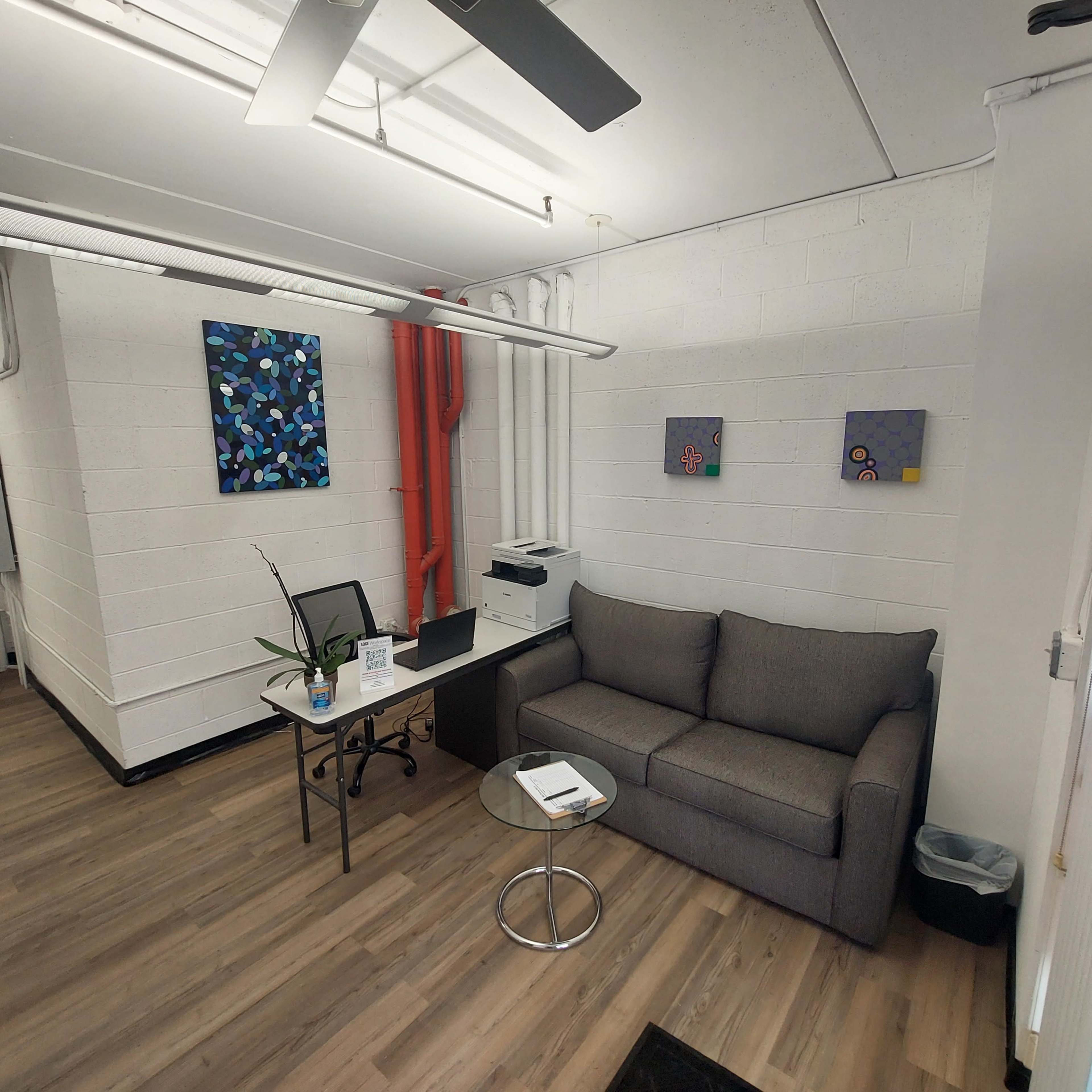 The image shows a small office space with a gray couch, a glass side table, a desk with a chair, and abstract paintings on the wall.