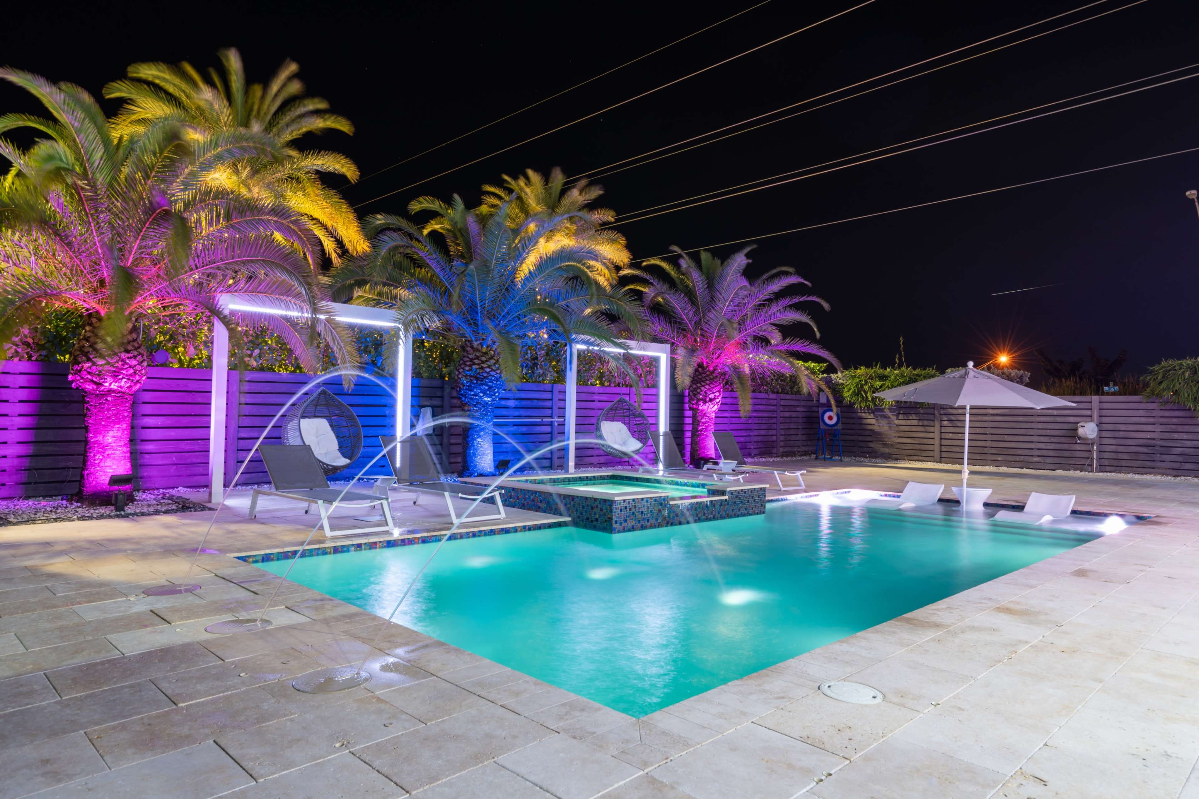 A modern pool area with colorful lighting, palm trees, and lounge chairs, set against a night sky.
