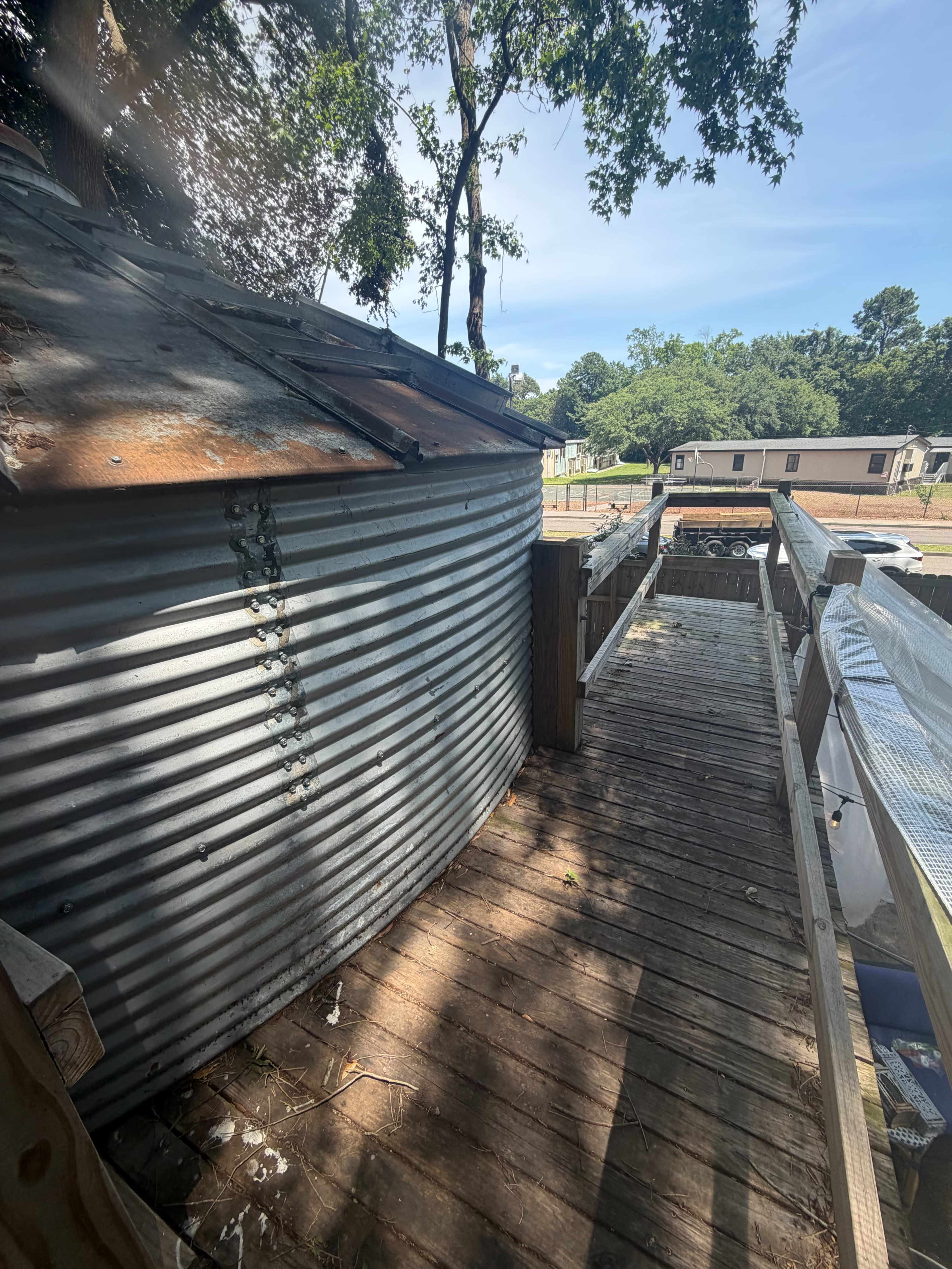 A rustic, corrugated metal structure stands beside a wooden walkway under a clear blue sky, with trees in the background.