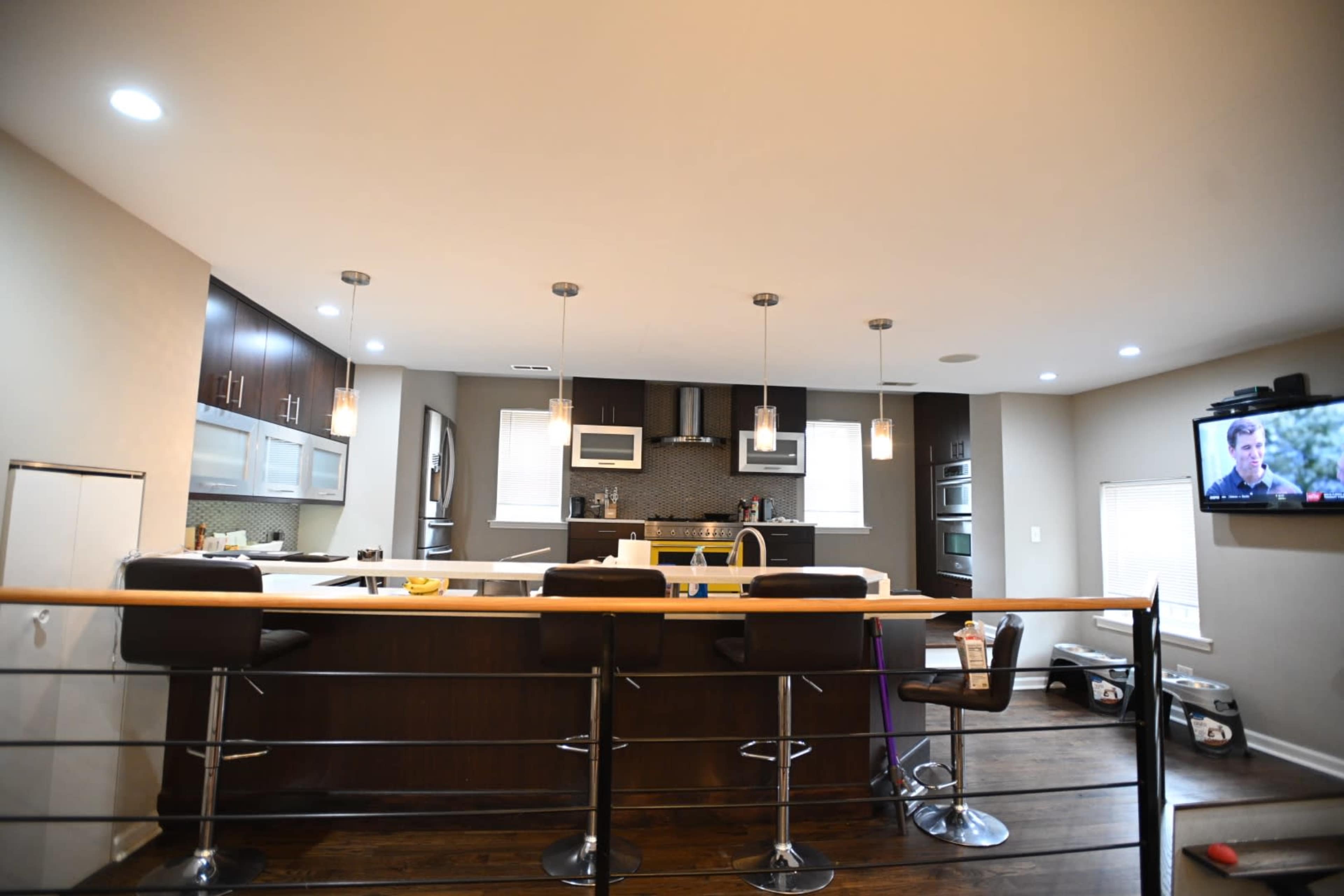 The kitchen features modern appliances, bar seating, and sleek cabinetry, with a living area visible in the background that includes a television.