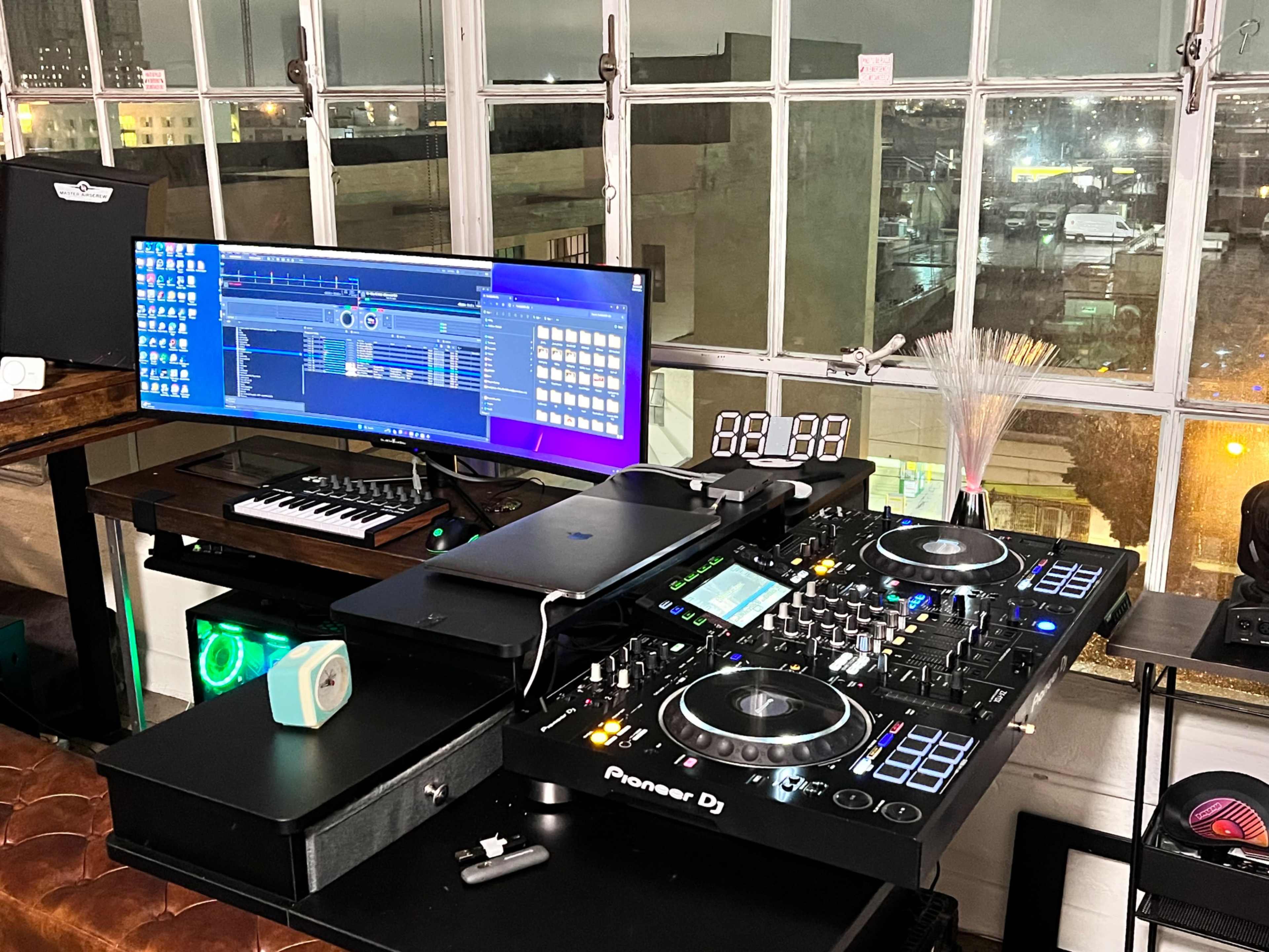 A DJ setup features a Pioneer DJ mixer, a laptop, a MIDI keyboard, and a large monitor displaying audio software, all positioned near a window overlooking a city skyline.