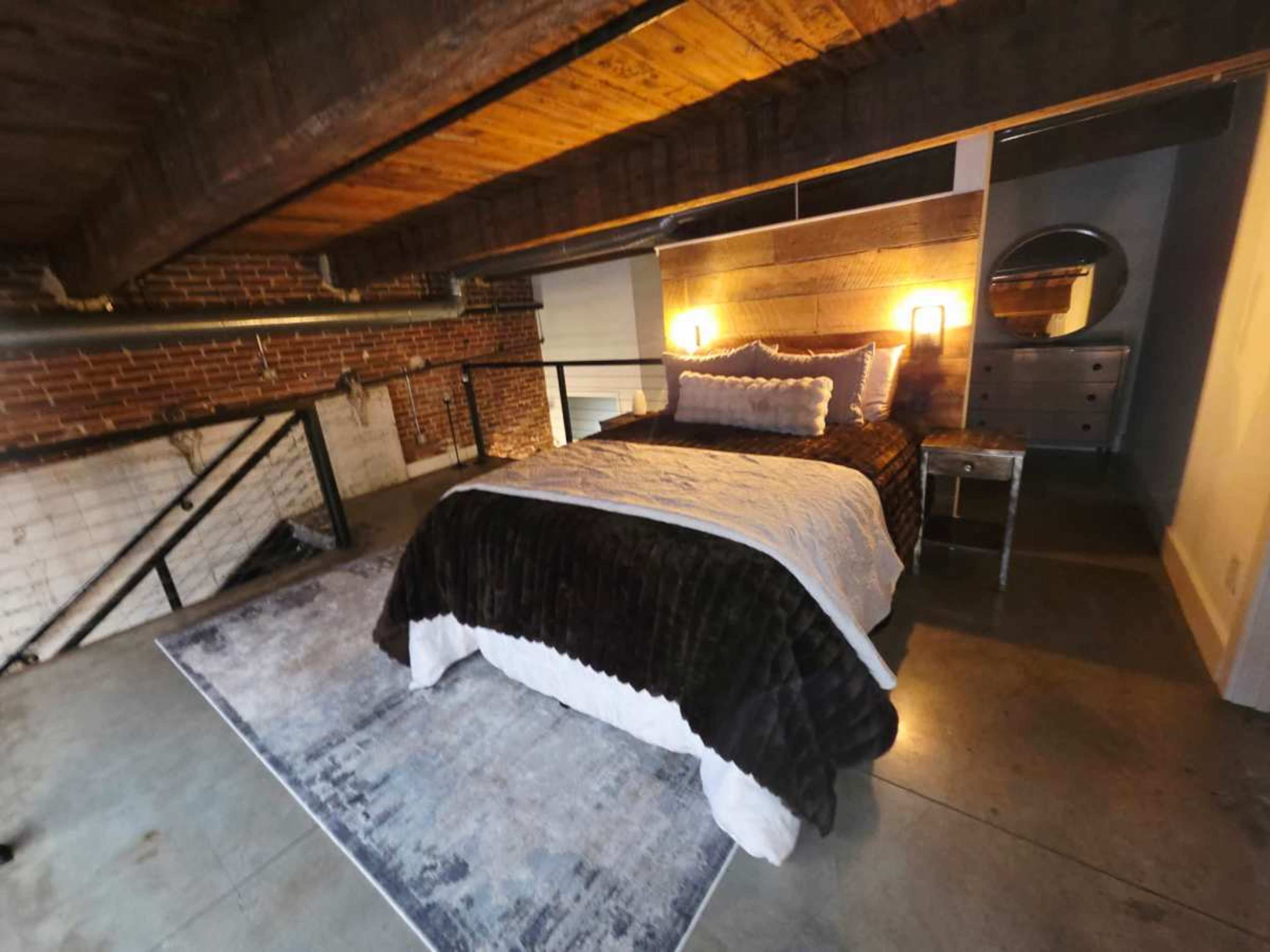 The image shows a modern bedroom with a dark wooden ceiling, exposed brick walls, and a bed surrounded by minimalist furniture.