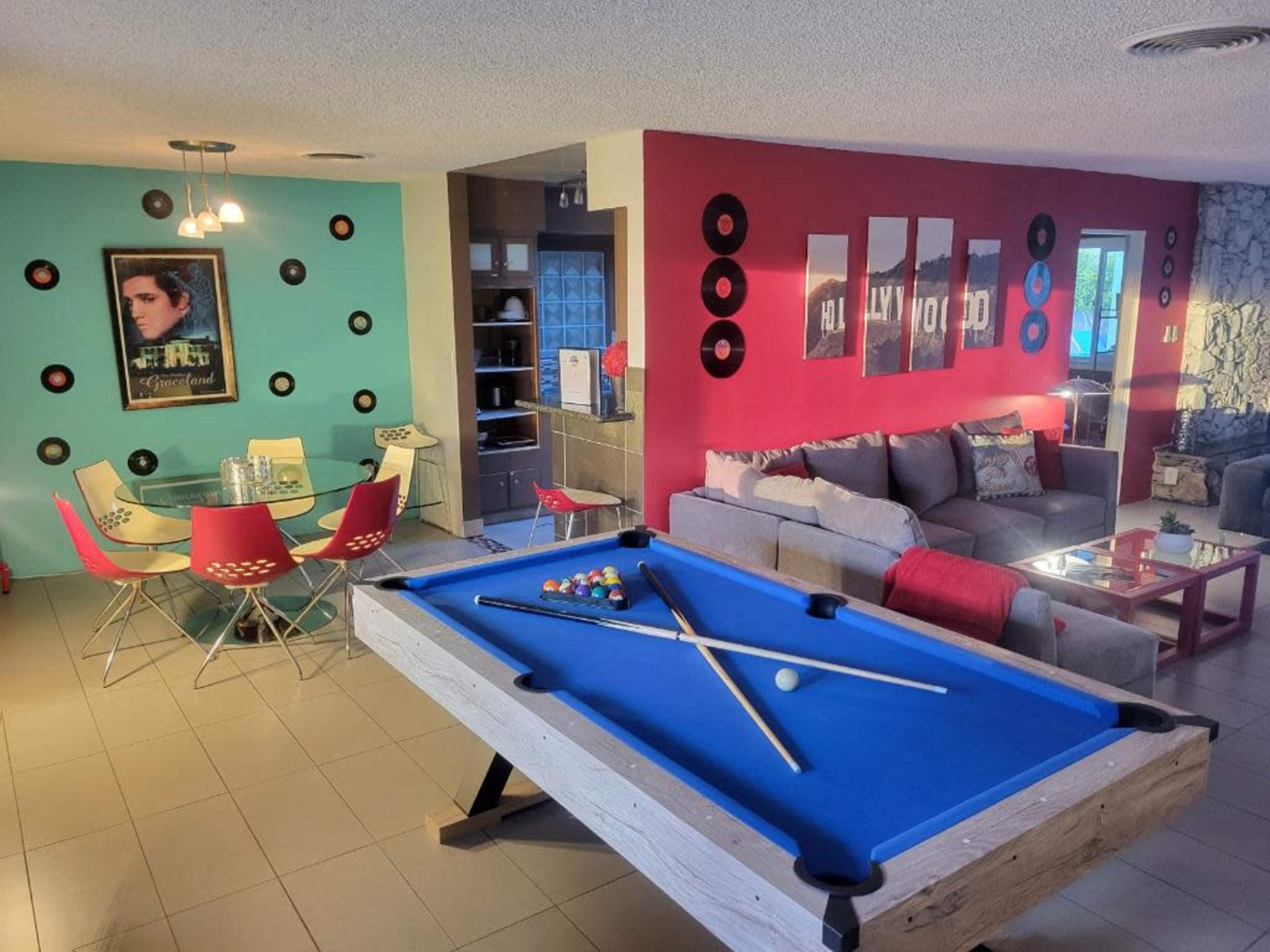A vibrant room featuring a pool table in the foreground, with a dining area and living space in the background, decorated with colorful artwork and furniture.