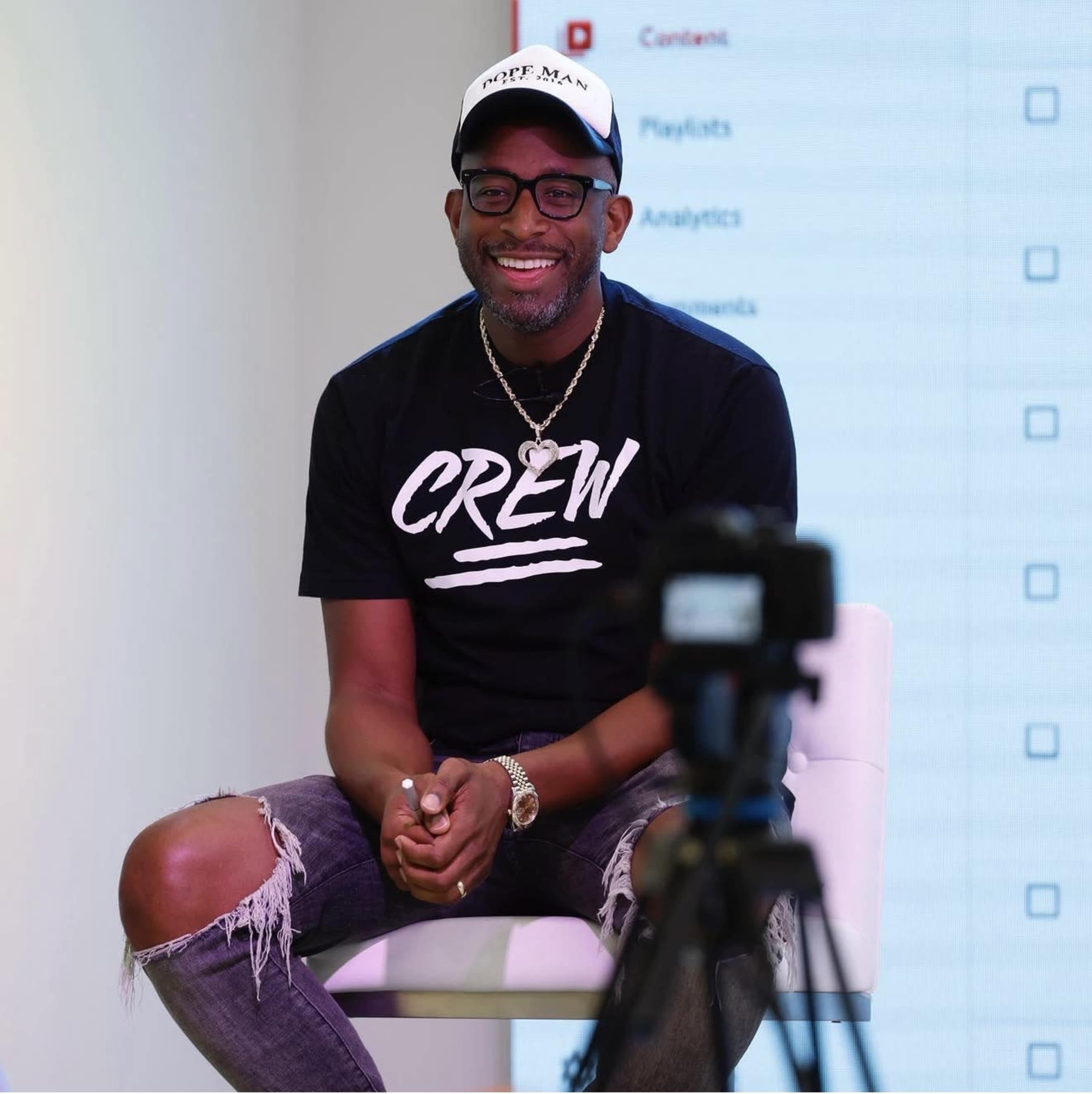A man wearing a black "CREW" T-shirt and a cap smiles while sitting on a white chair in front of a camera and a screen displaying a checklist.