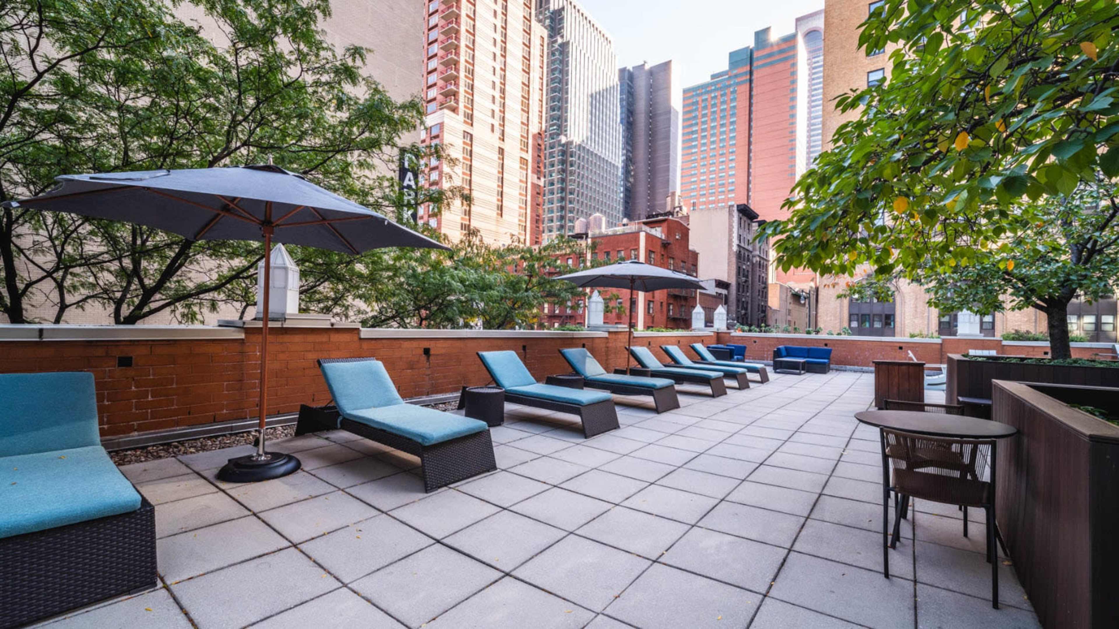 A rooftop patio with lounge chairs, an umbrella, and surrounding buildings in an urban setting.