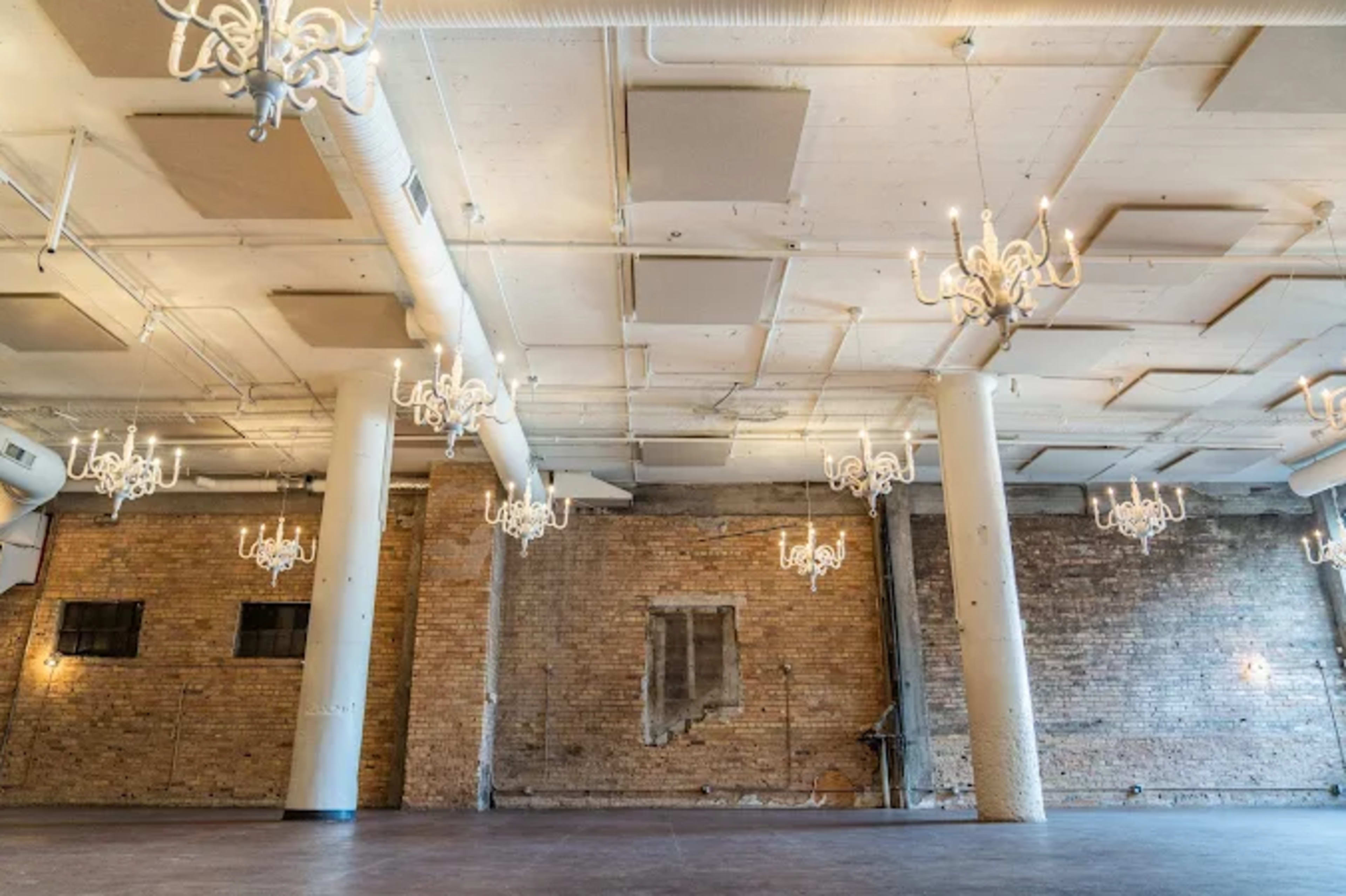 The image shows a large, empty room with exposed brick walls, white pillars, and multiple chandeliers hanging from the ceiling.