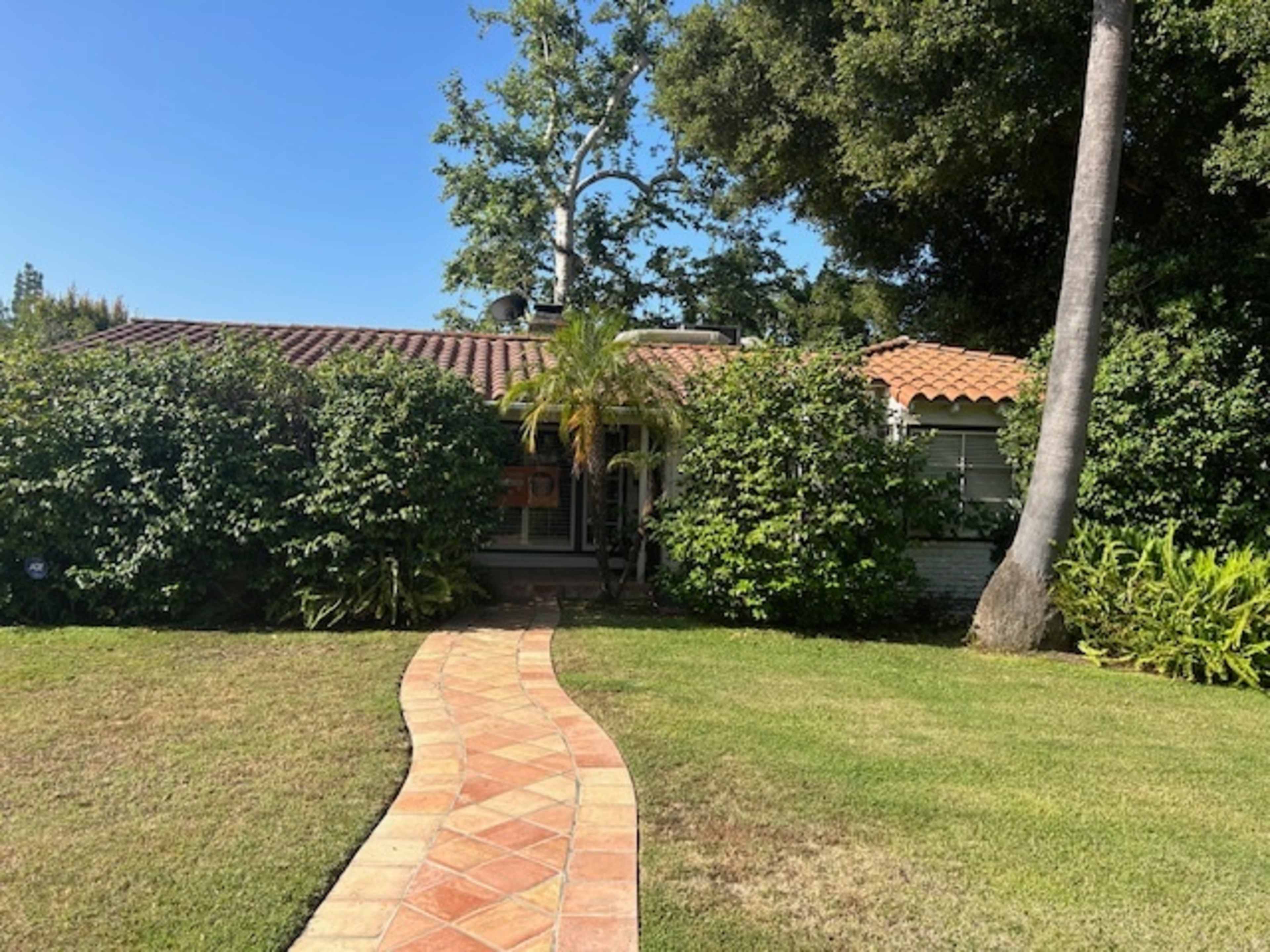 A brick pathway leads through a landscaped yard to a single-story house with a tiled roof and surrounding greenery.
