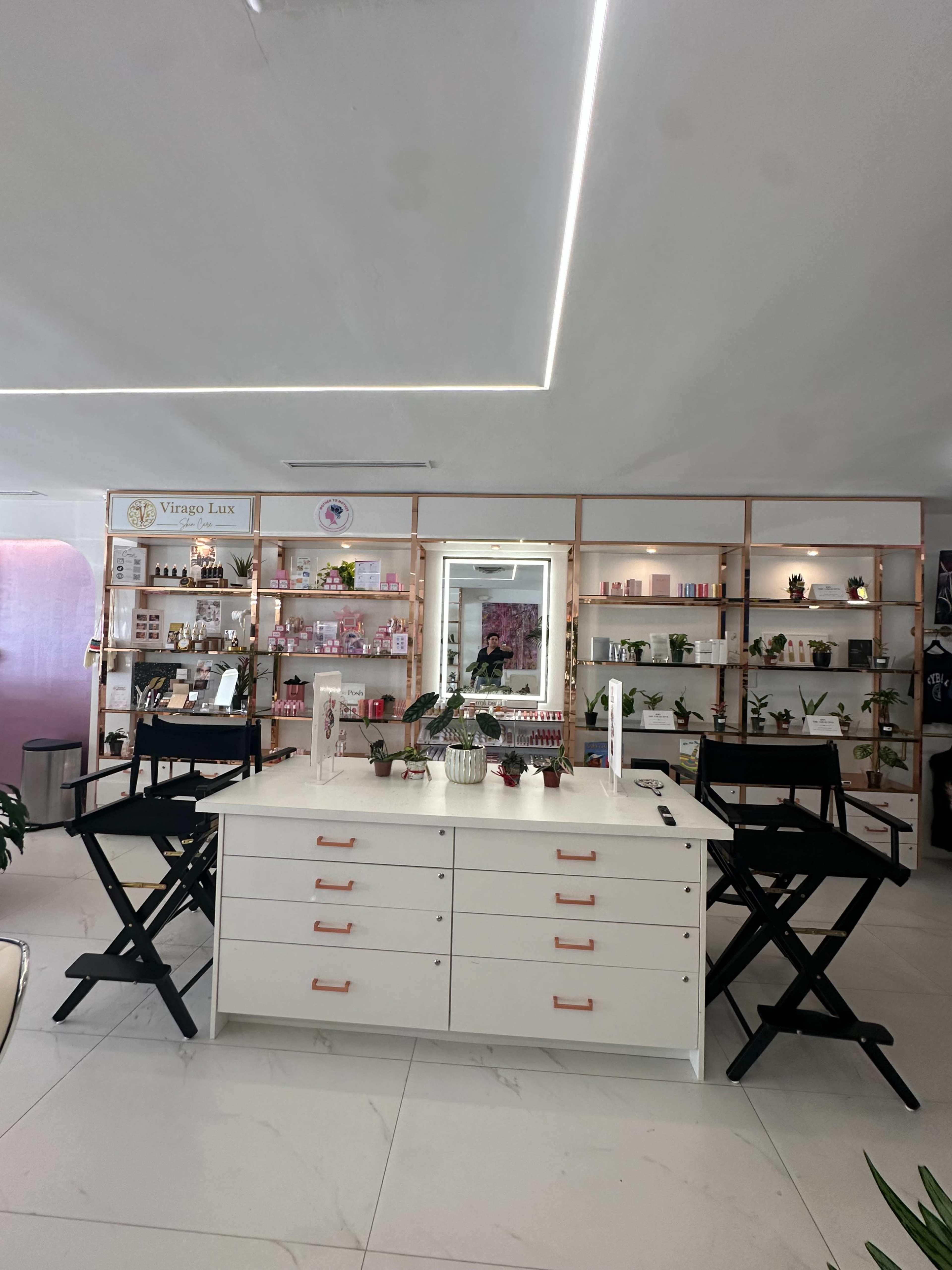 The image shows a modern beauty studio with neatly arranged shelves of products, a large mirror, and two black director's chairs in front of a white cabinet.