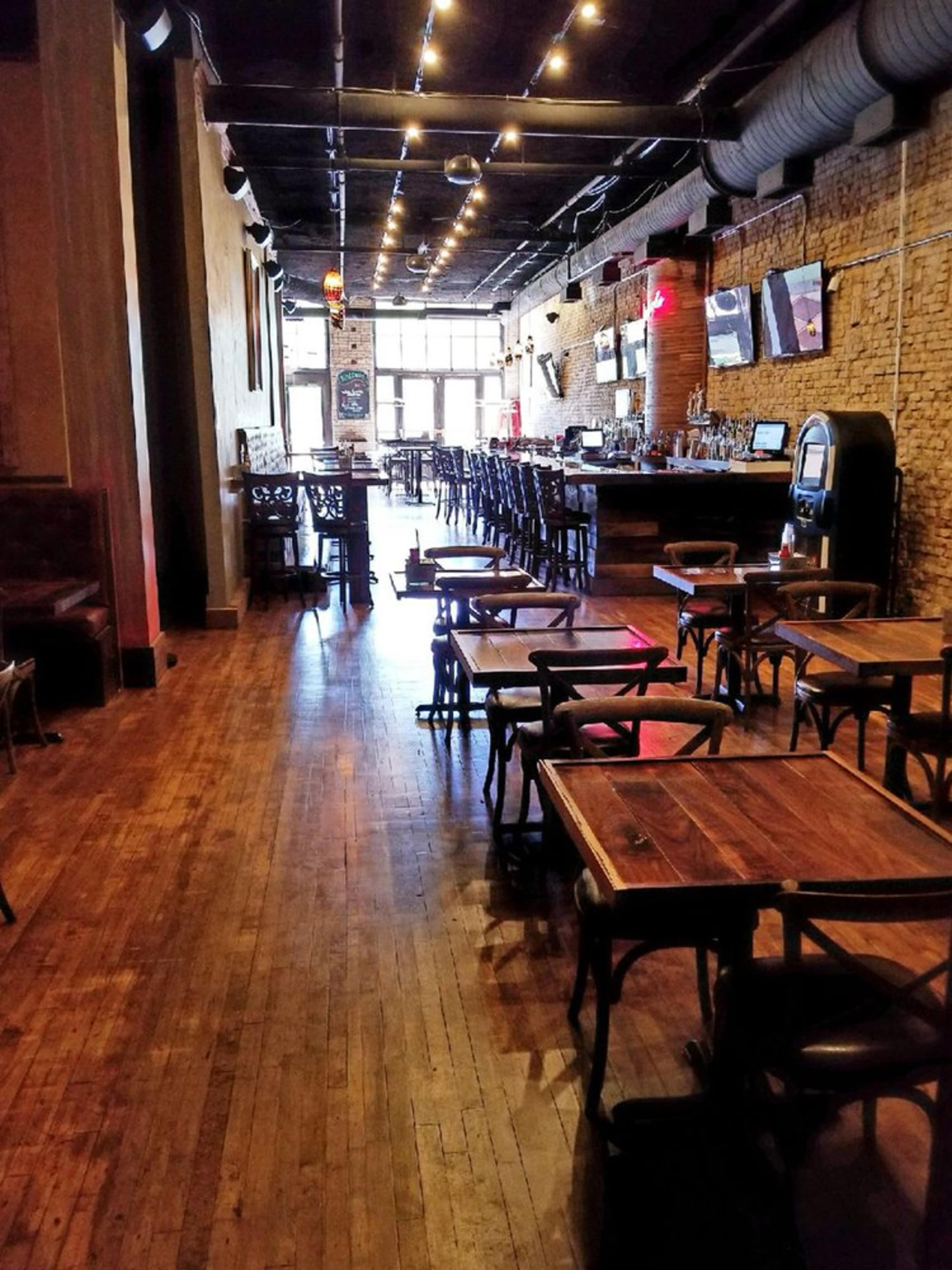 A dimly lit bar interior with wooden floors, empty tables, and a bar counter at the back.