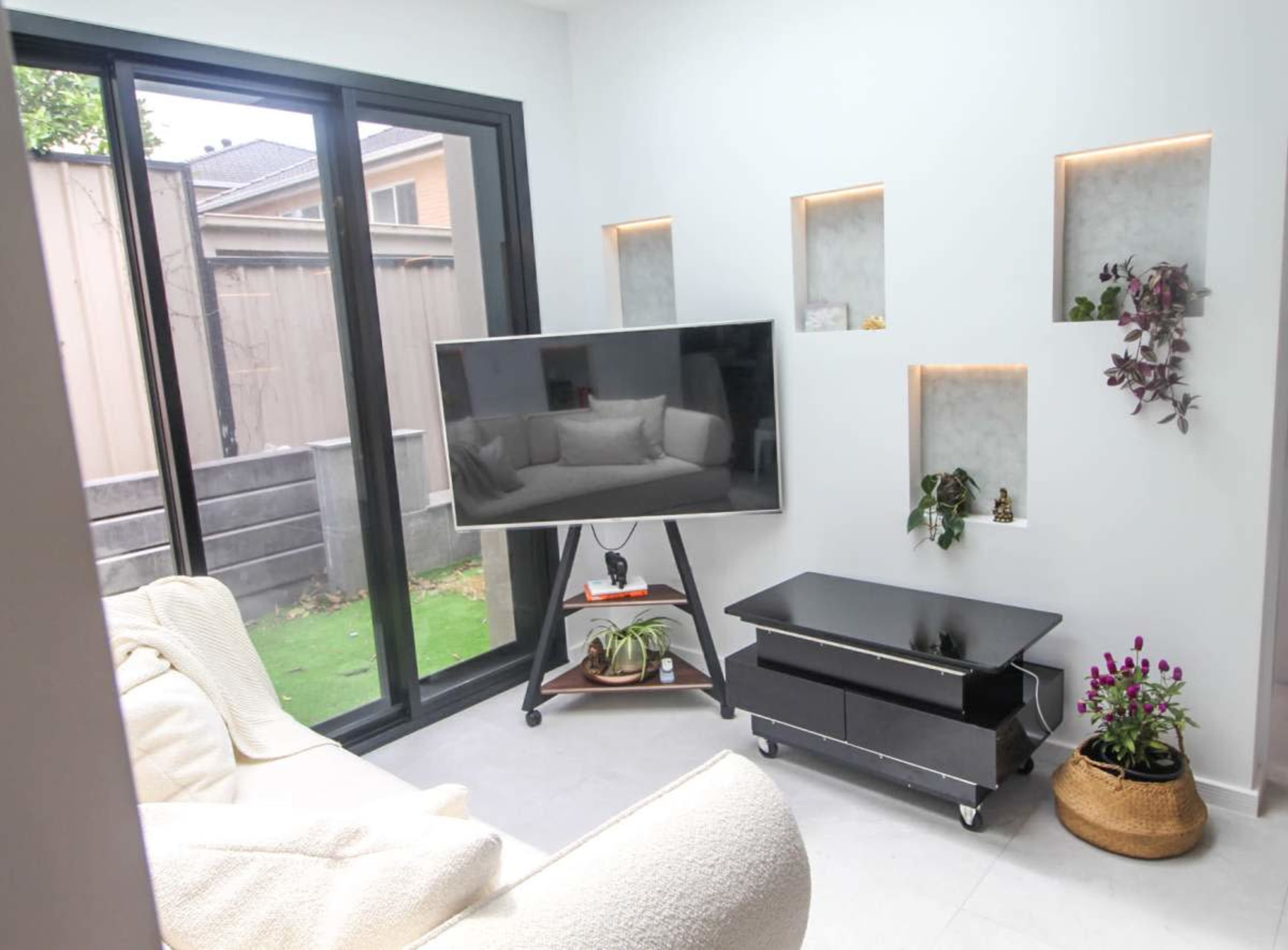 The image shows a modern living room with a large television on a stand, a white sofa, and decorative niches with plants on the walls.