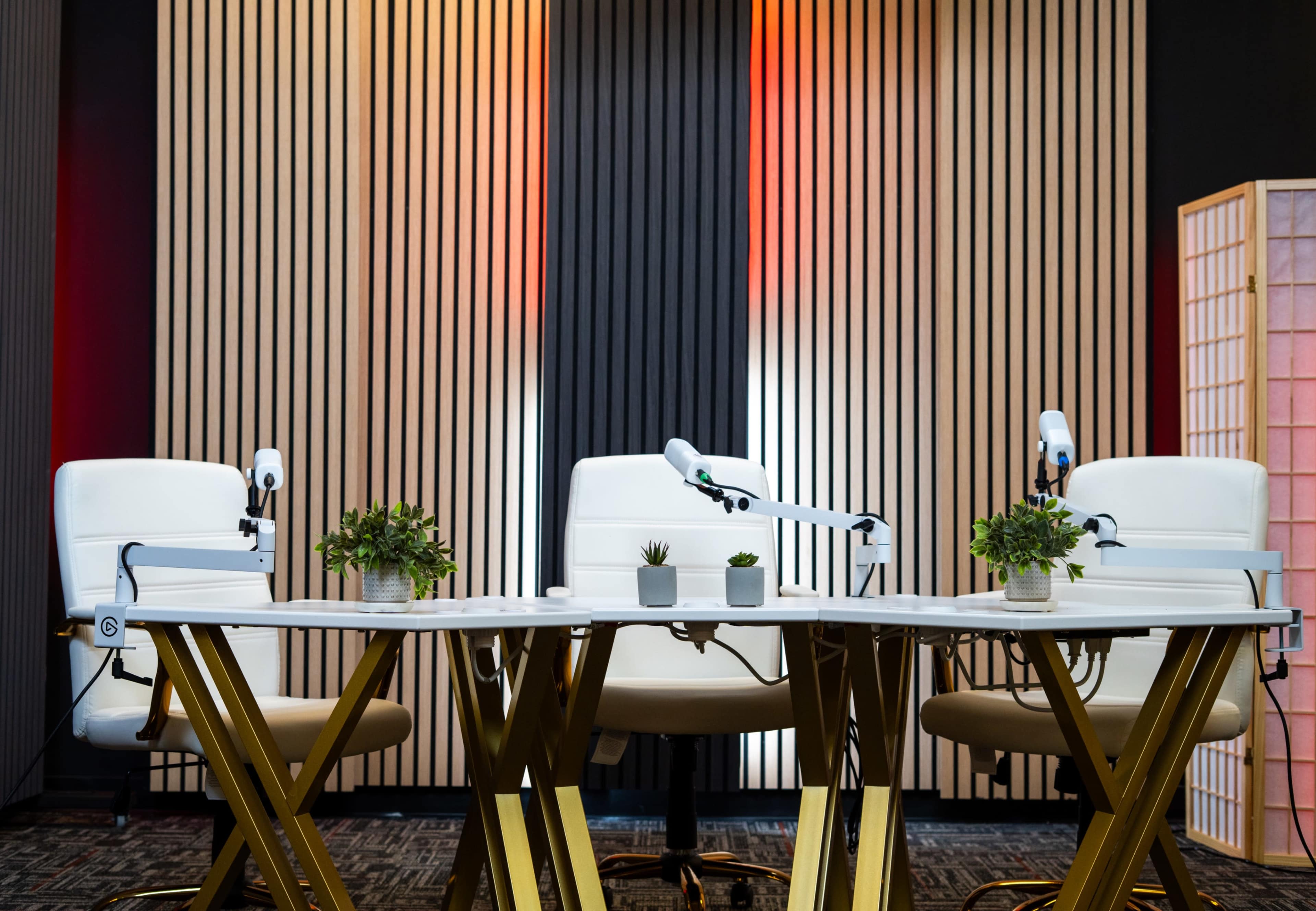 A modern podcast or interview setup features three white chairs with microphones arranged around a sleek table, accompanied by small potted plants against a backdrop of striped wood paneling.