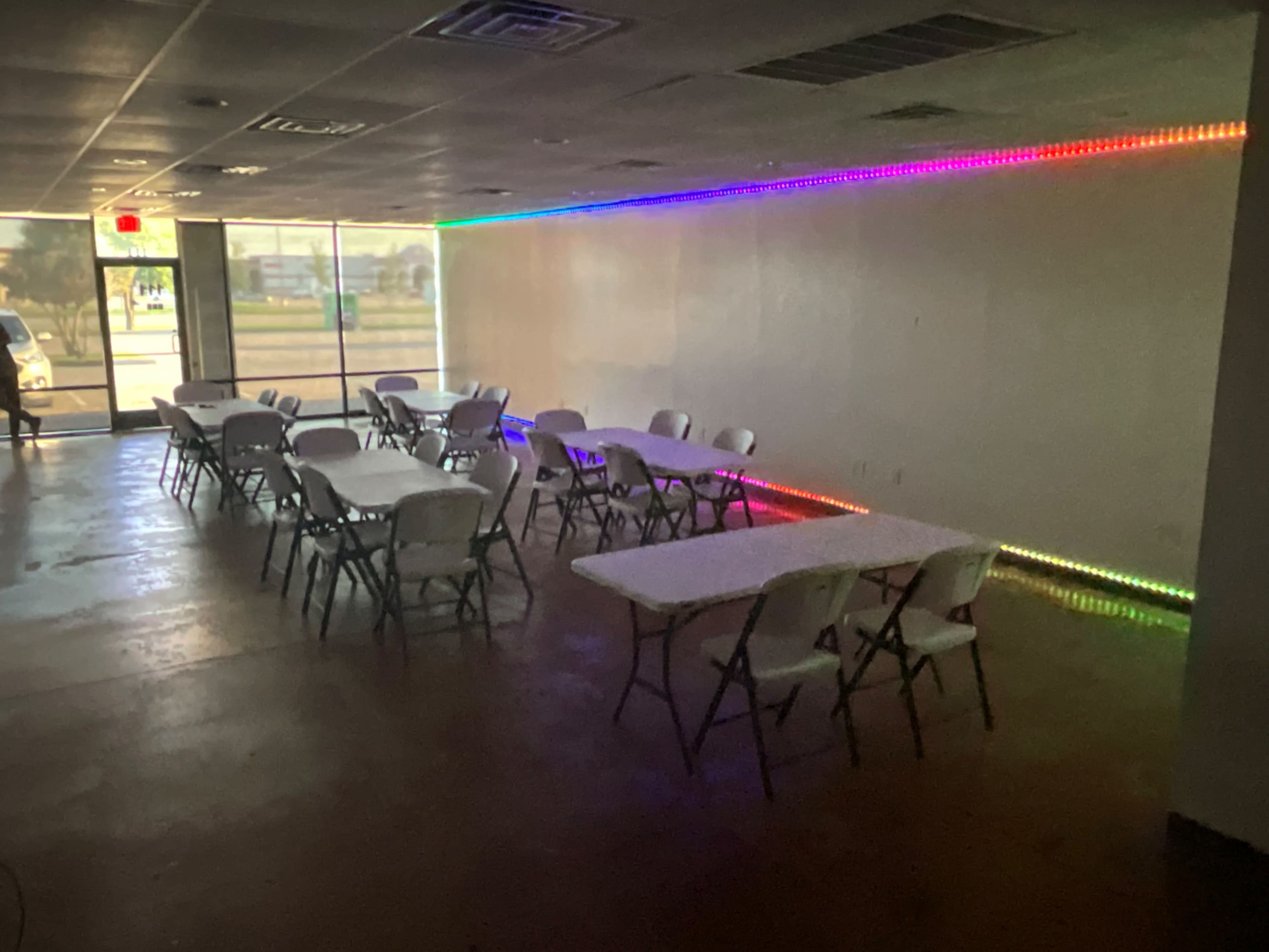The image shows an empty room with several white tables arranged in rows and a strip of colorful LED lights along one wall.