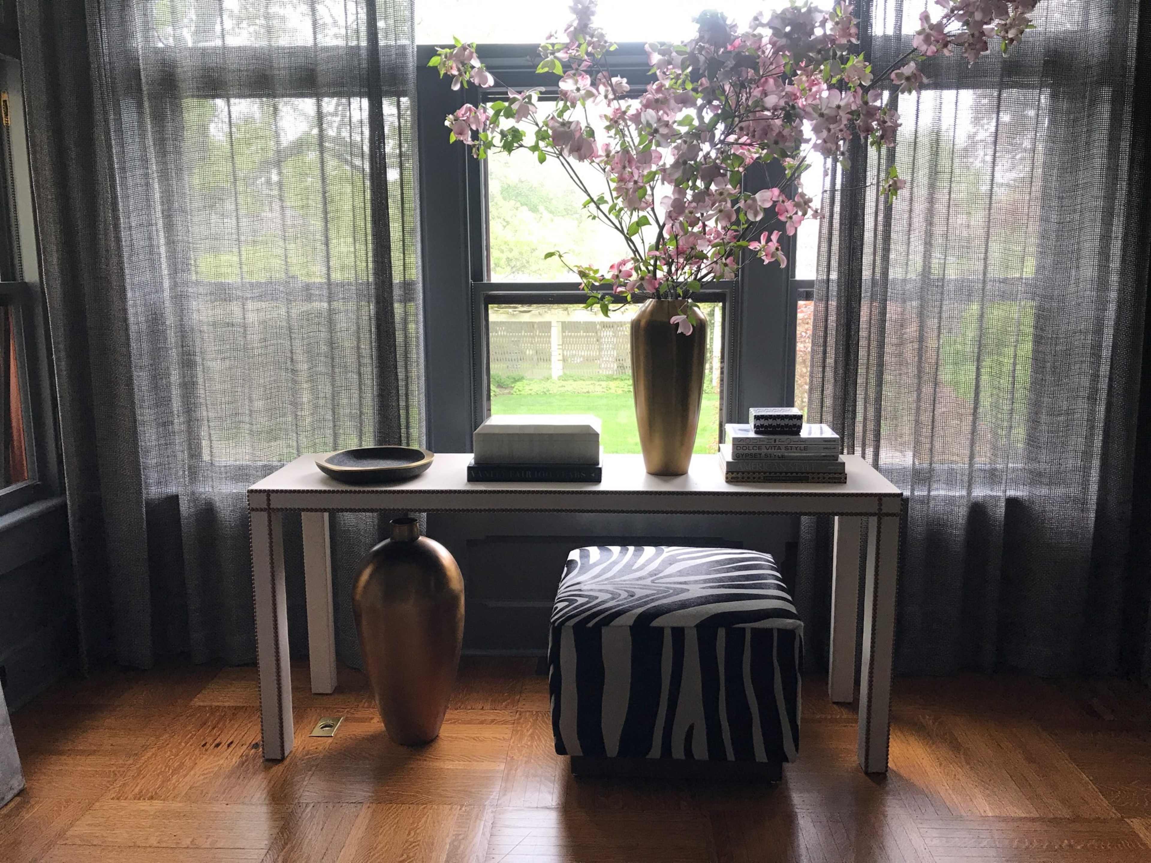 A table with a vase of flowers sits beside a decorative stool and books in front of a large window with sheer curtains.