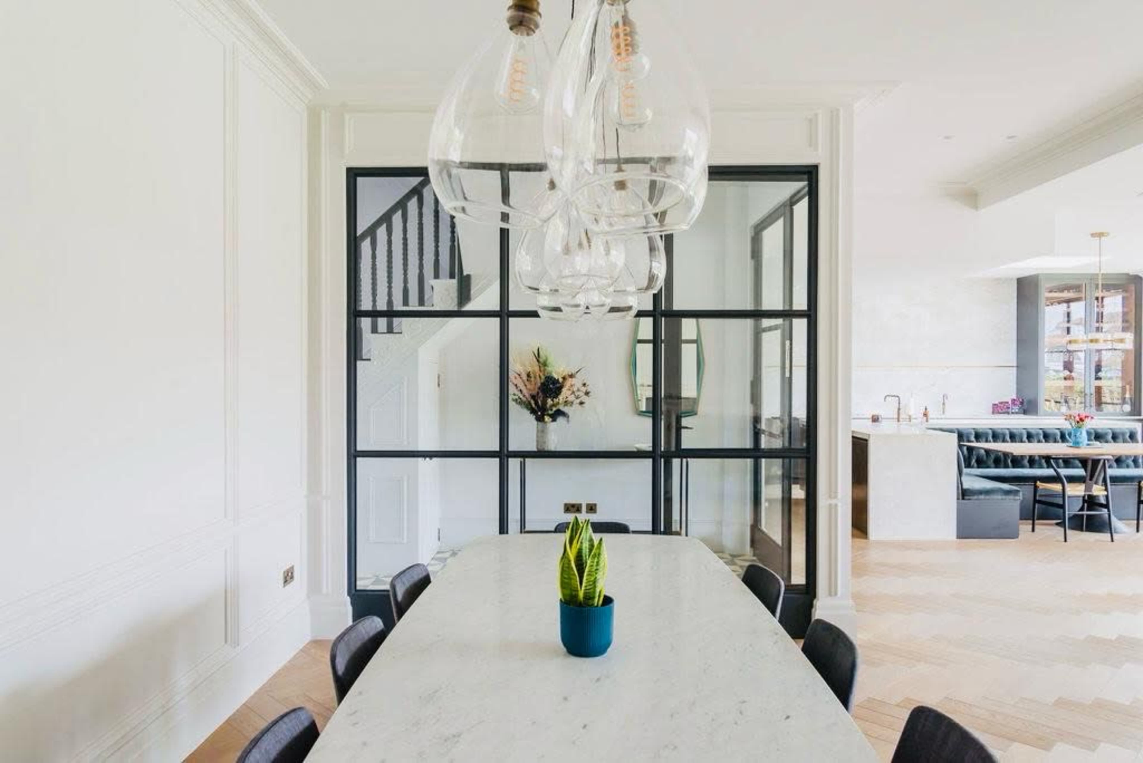 A long marble dining table is set under a cluster of glass pendant lights, with a green plant in a blue pot at its center, surrounded by black chairs in a bright, modern dining area.