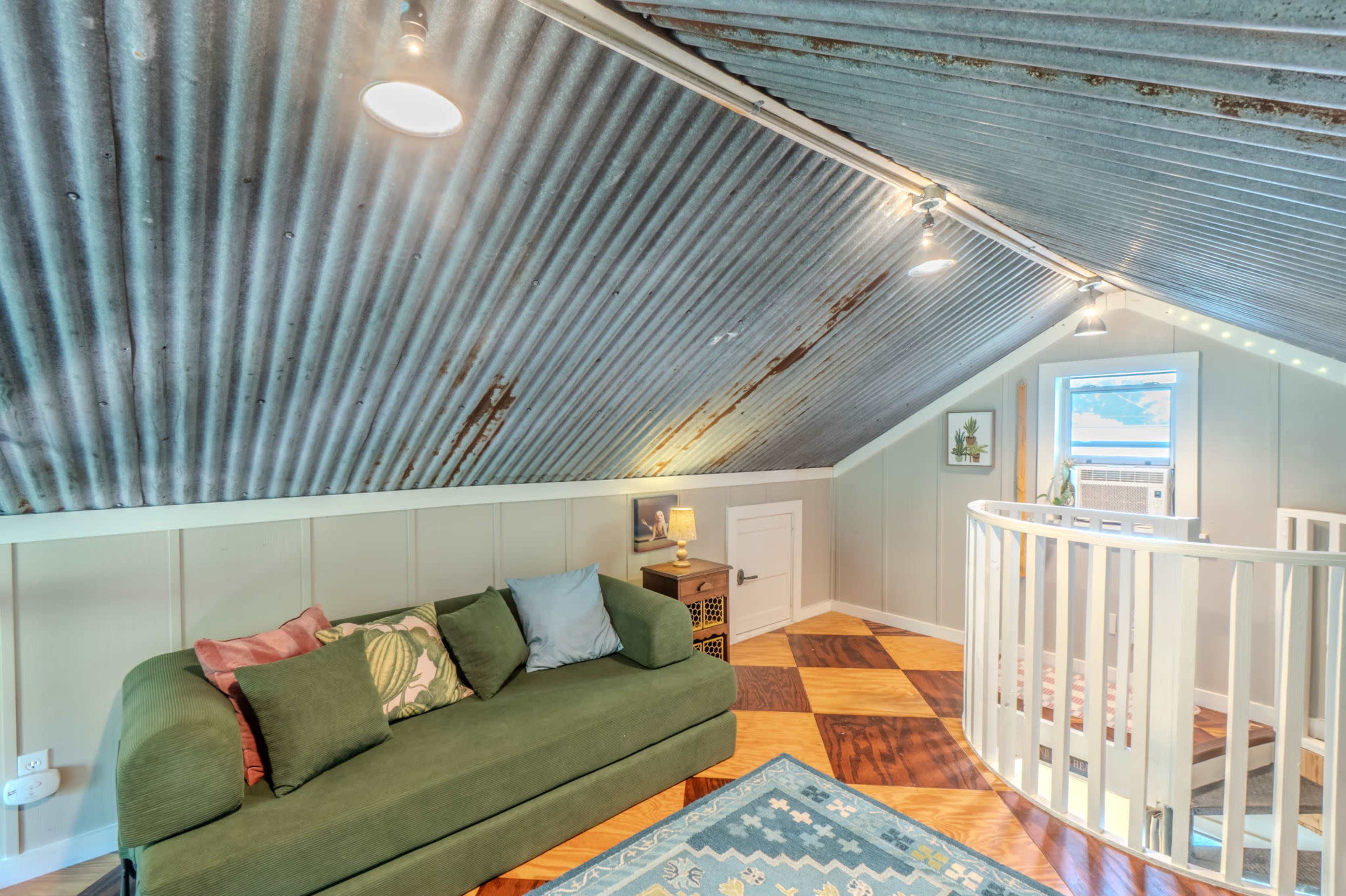 The image showcases a cozy loft space with a corrugated metal ceiling, a green sofa, wooden flooring, and a circular railing.