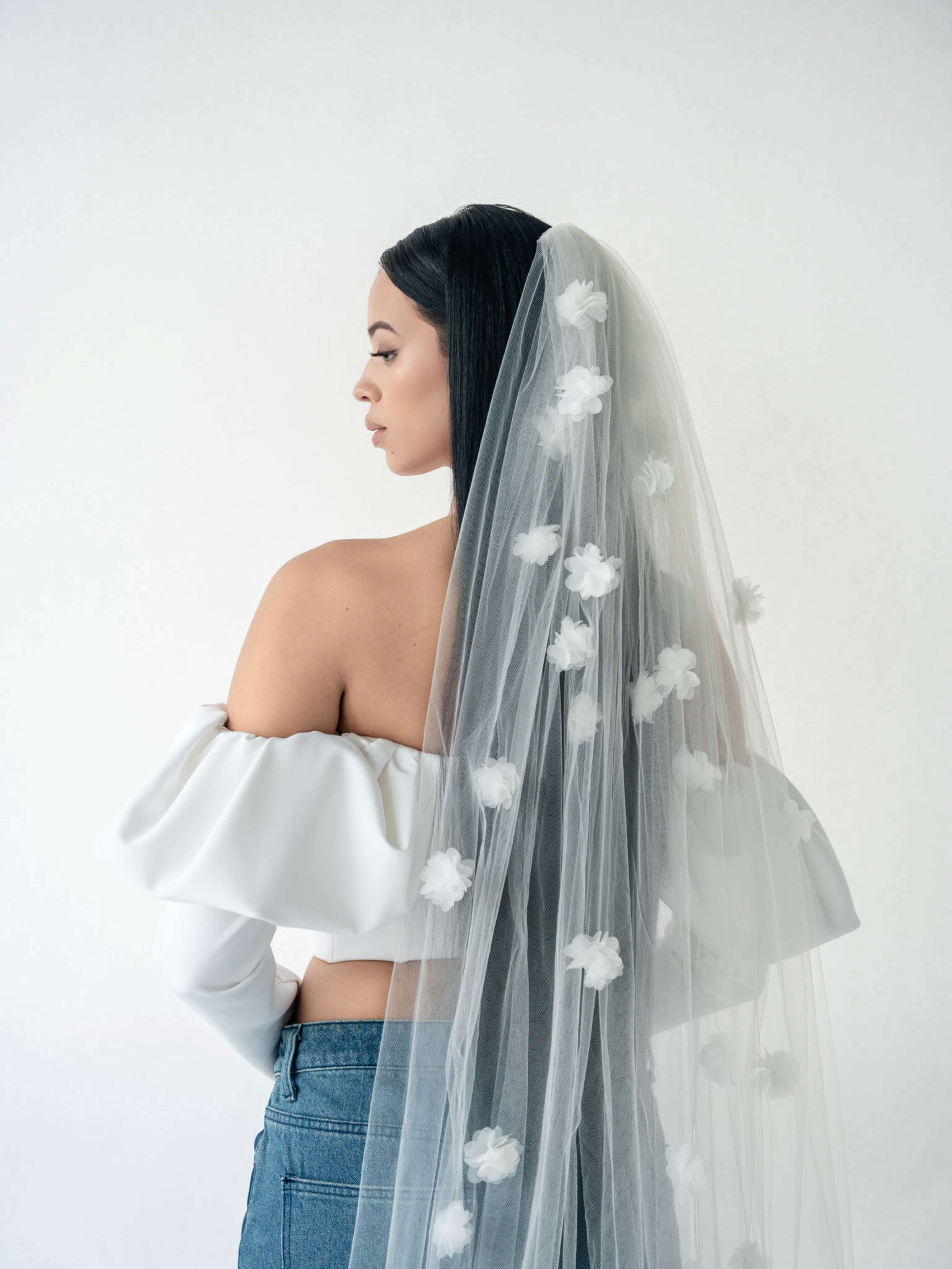 A woman with long black hair wears a white off-the-shoulder top and a sheer veil decorated with white flowers.