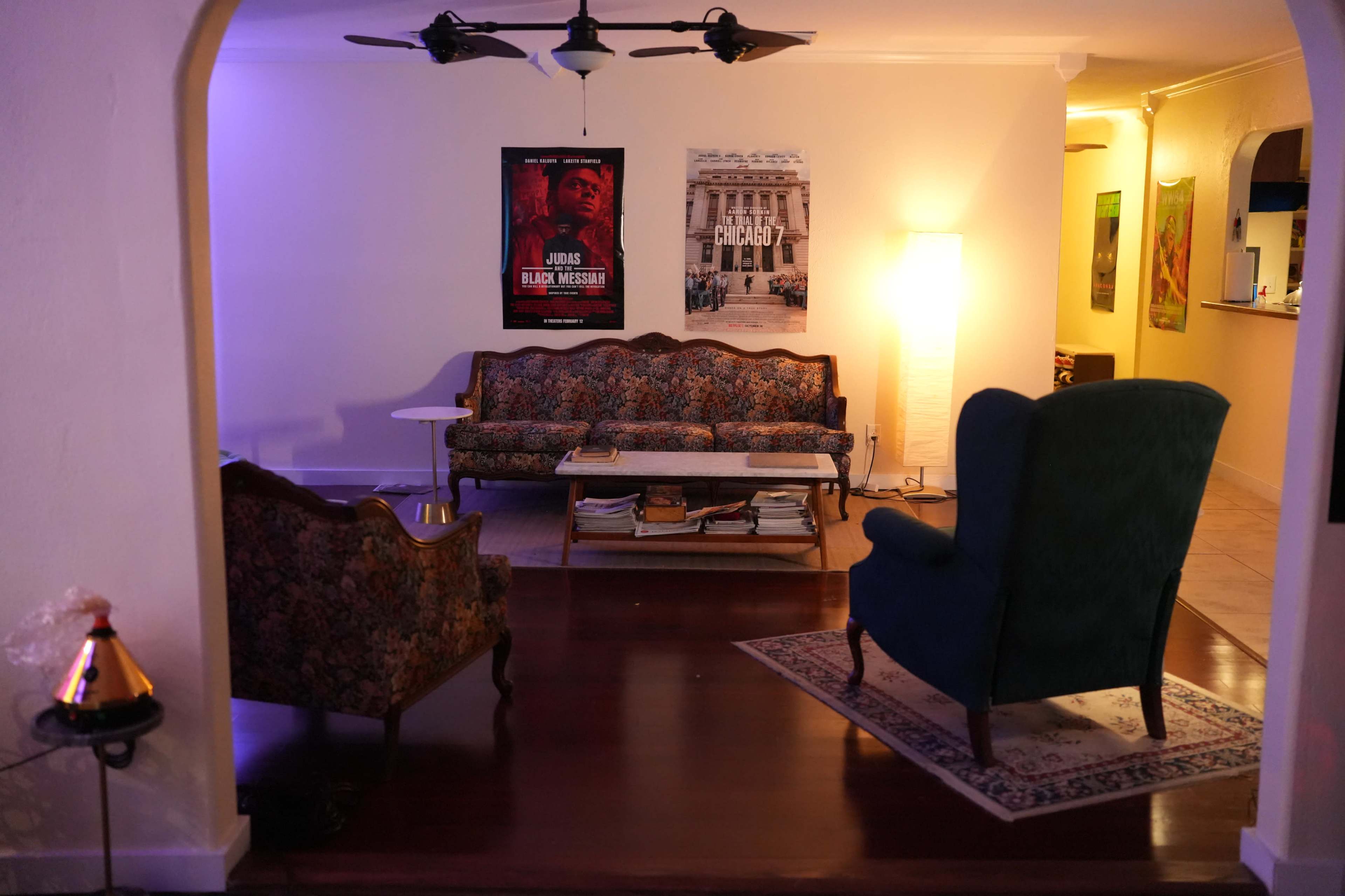 The image shows a living room featuring a patterned couch, two chairs, a coffee table with stacked books, and wall posters in a softly lit space.