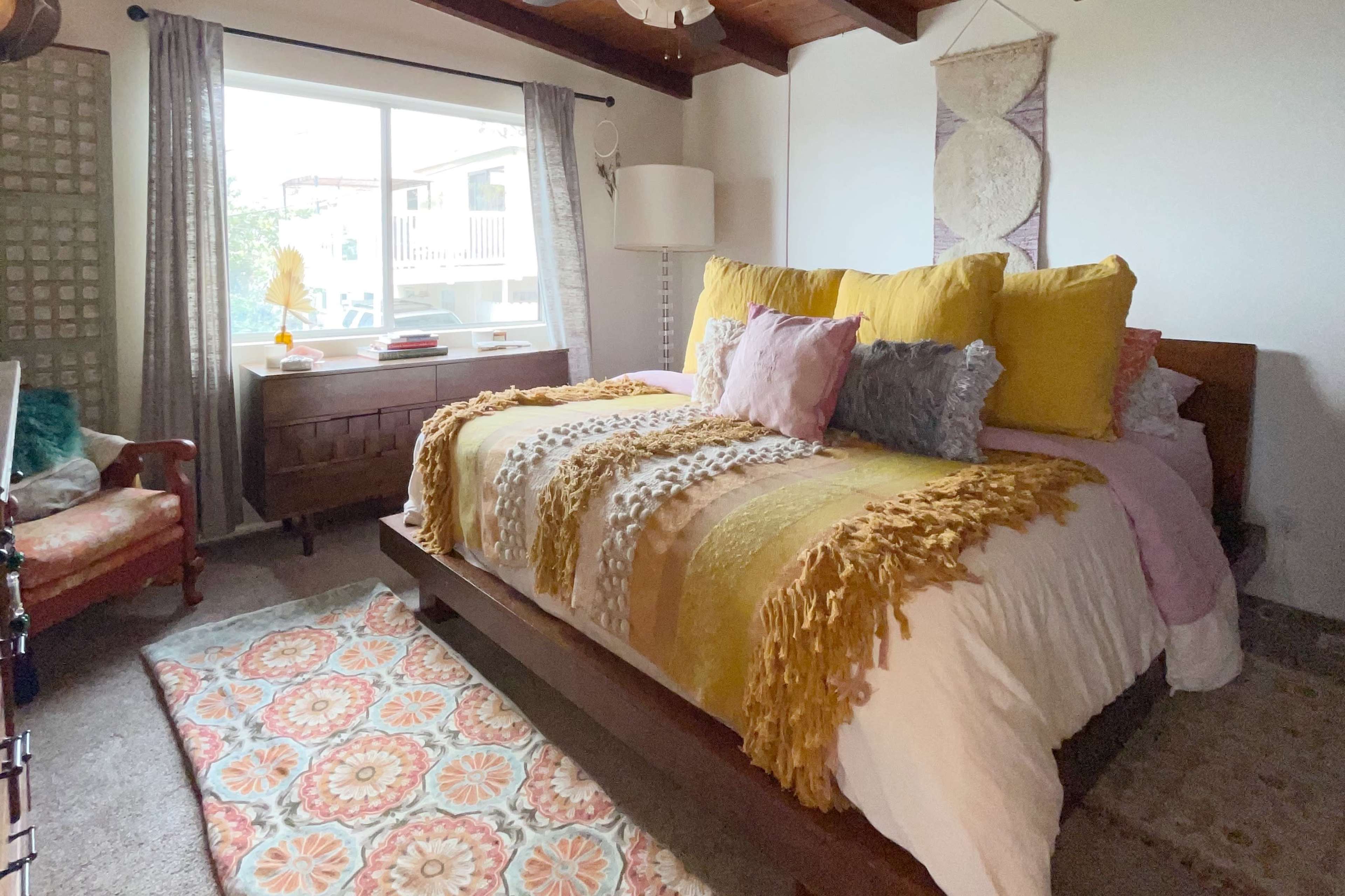 A cozy bedroom features a large bed with colorful pillows, a patterned rug, and a window overlooking an outdoor area.