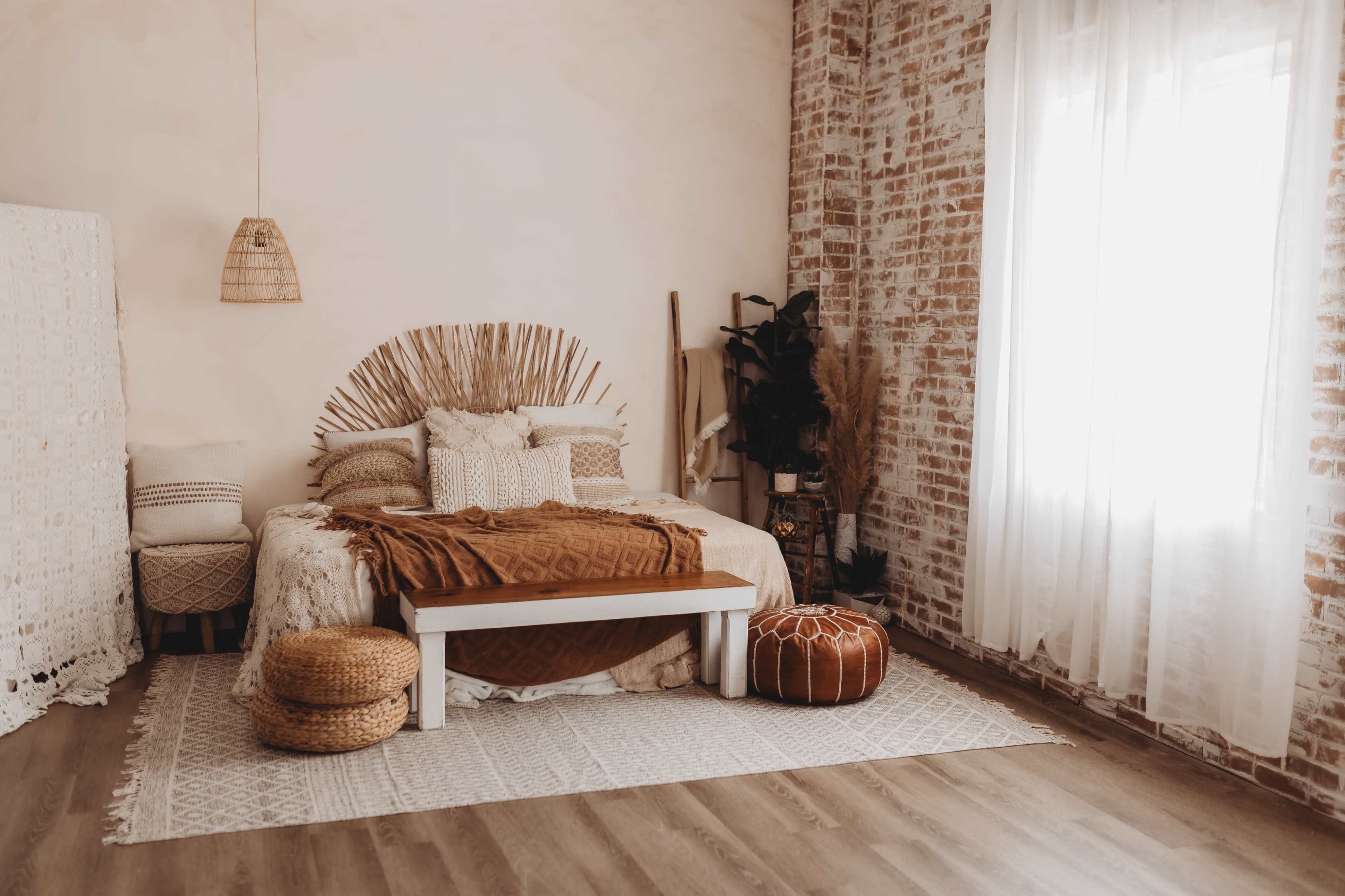 A cozy bedroom featuring a bed with a woven headboard, a large blanket, and natural decor elements, set against a backdrop of exposed brick walls and a bright window.