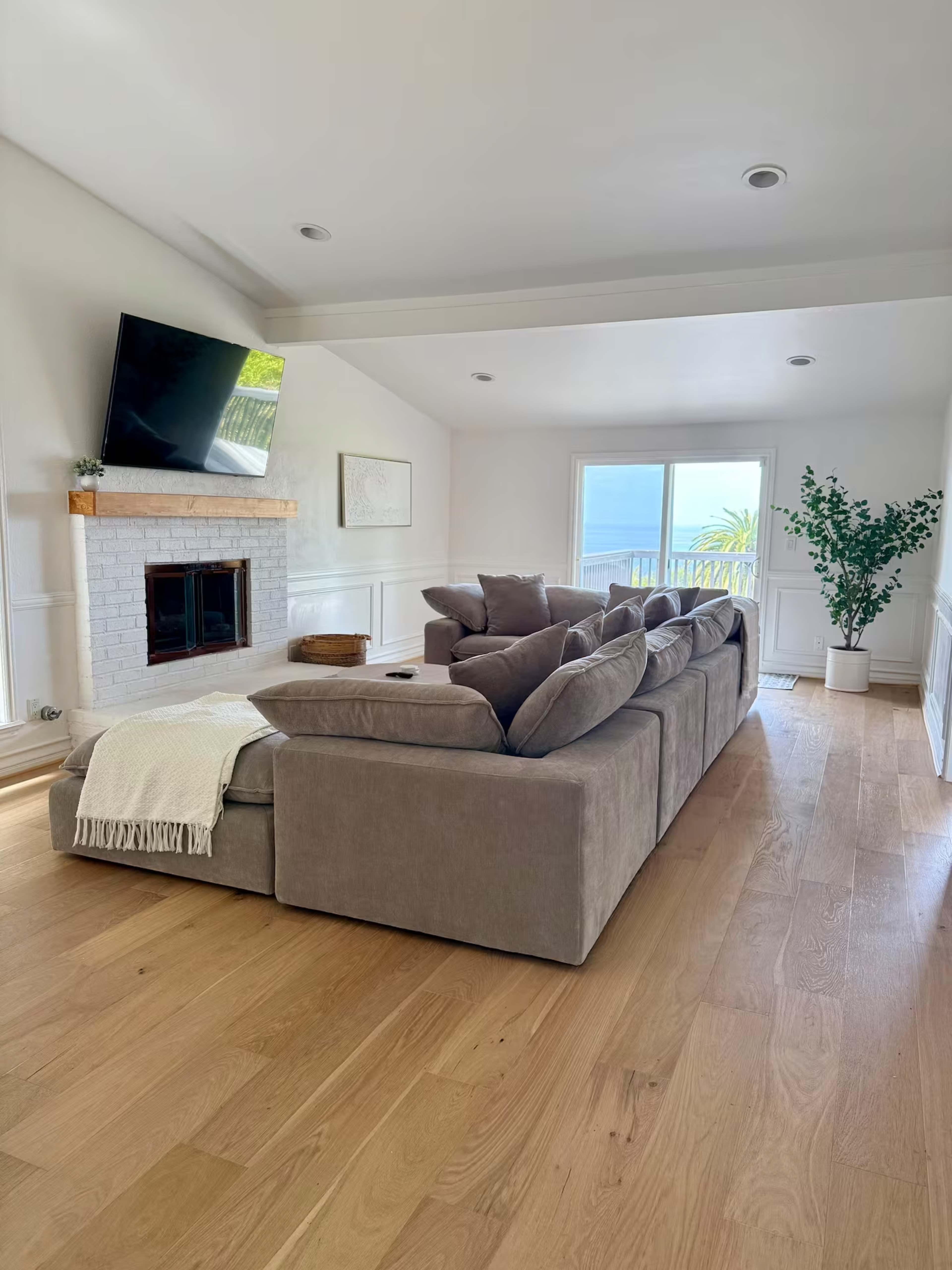 The image shows a modern living room featuring a large sectional sofa, a fireplace with a wooden mantel, and a view of the outdoors through large windows.