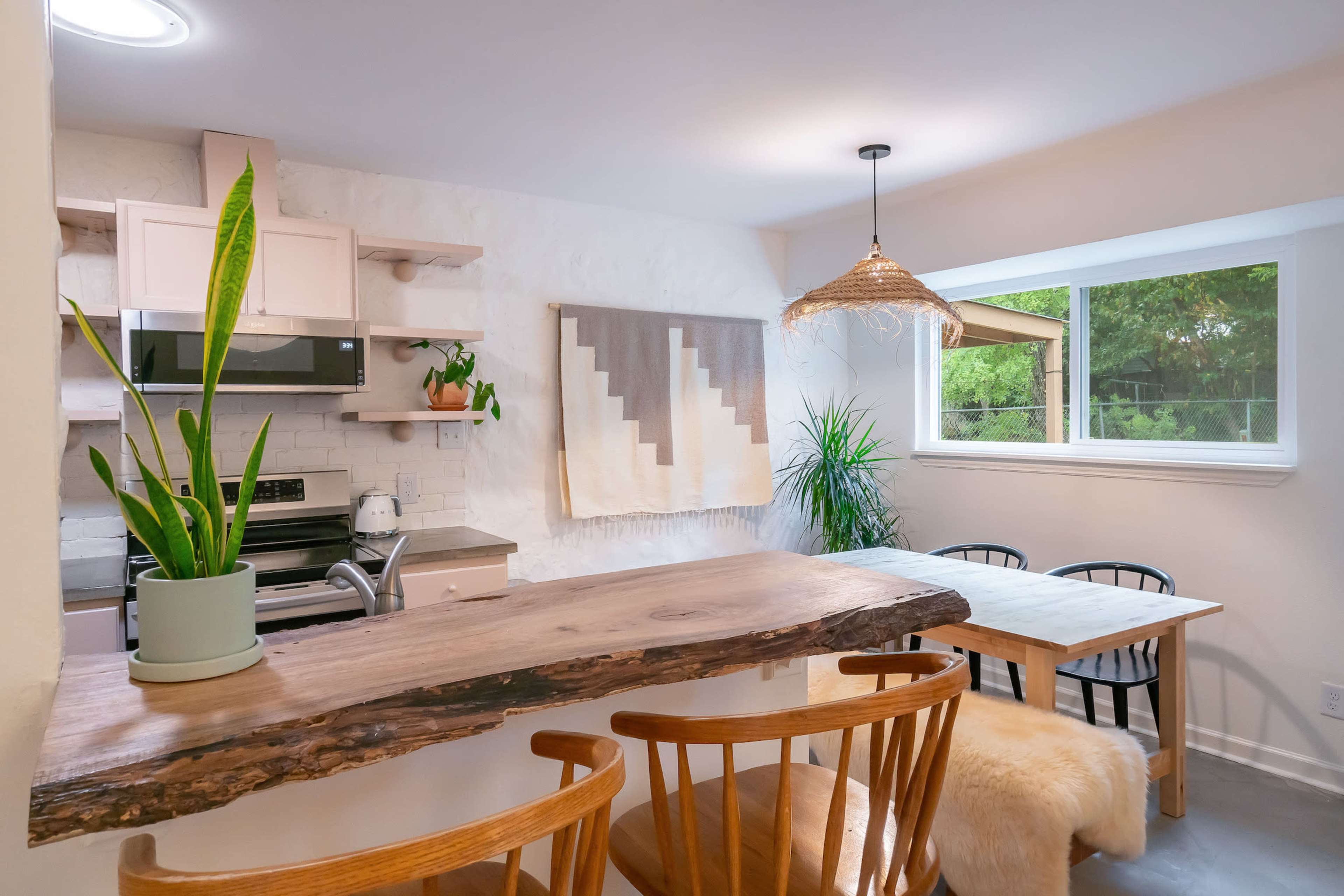 A modern kitchen features a wooden dining table, minimalist decor, and plants, with natural light coming through a window.