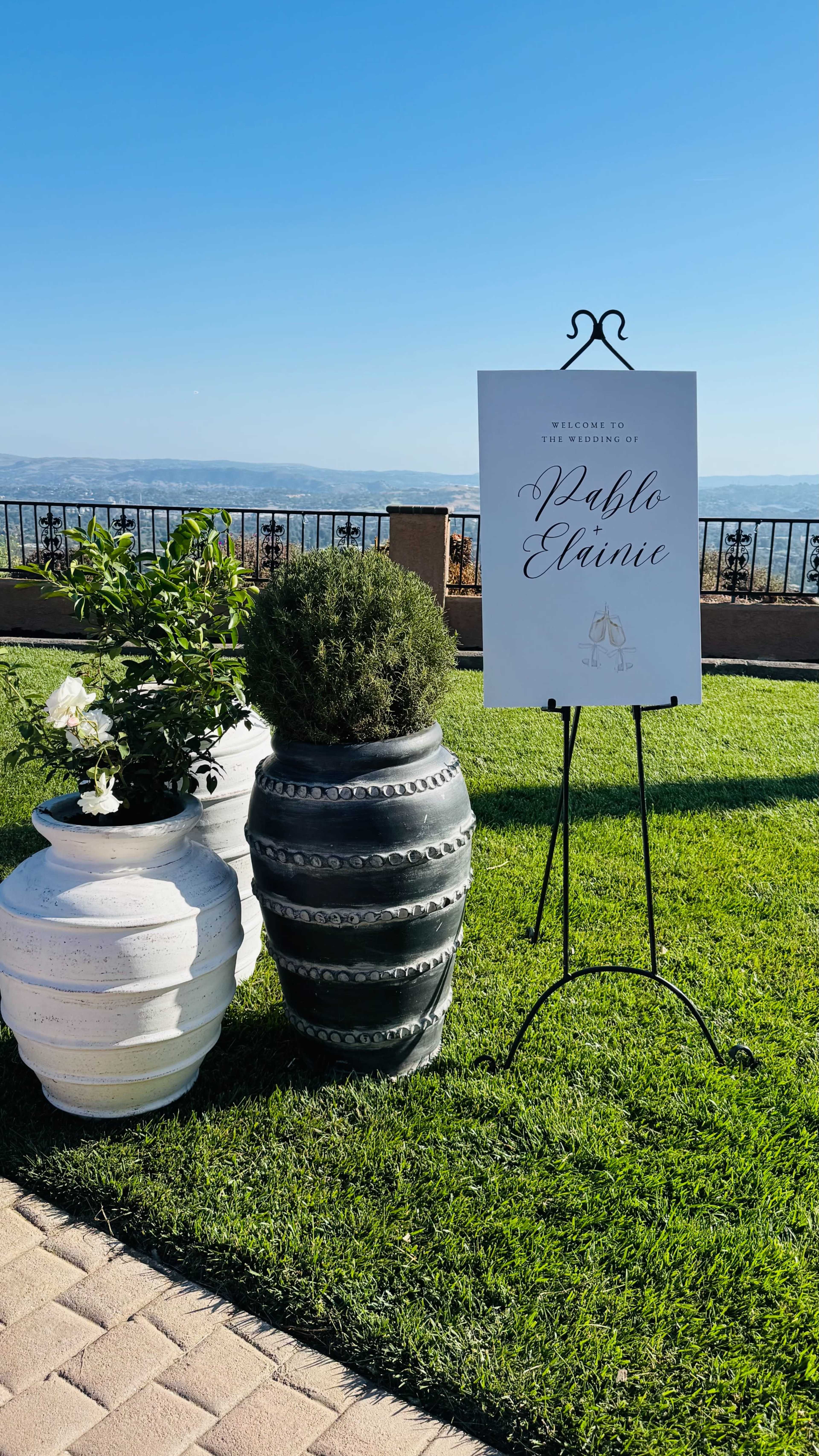 A sign reading "Welcome to Pablo & Elaine" stands beside decorative planters on a green lawn with a clear blue sky in the background.