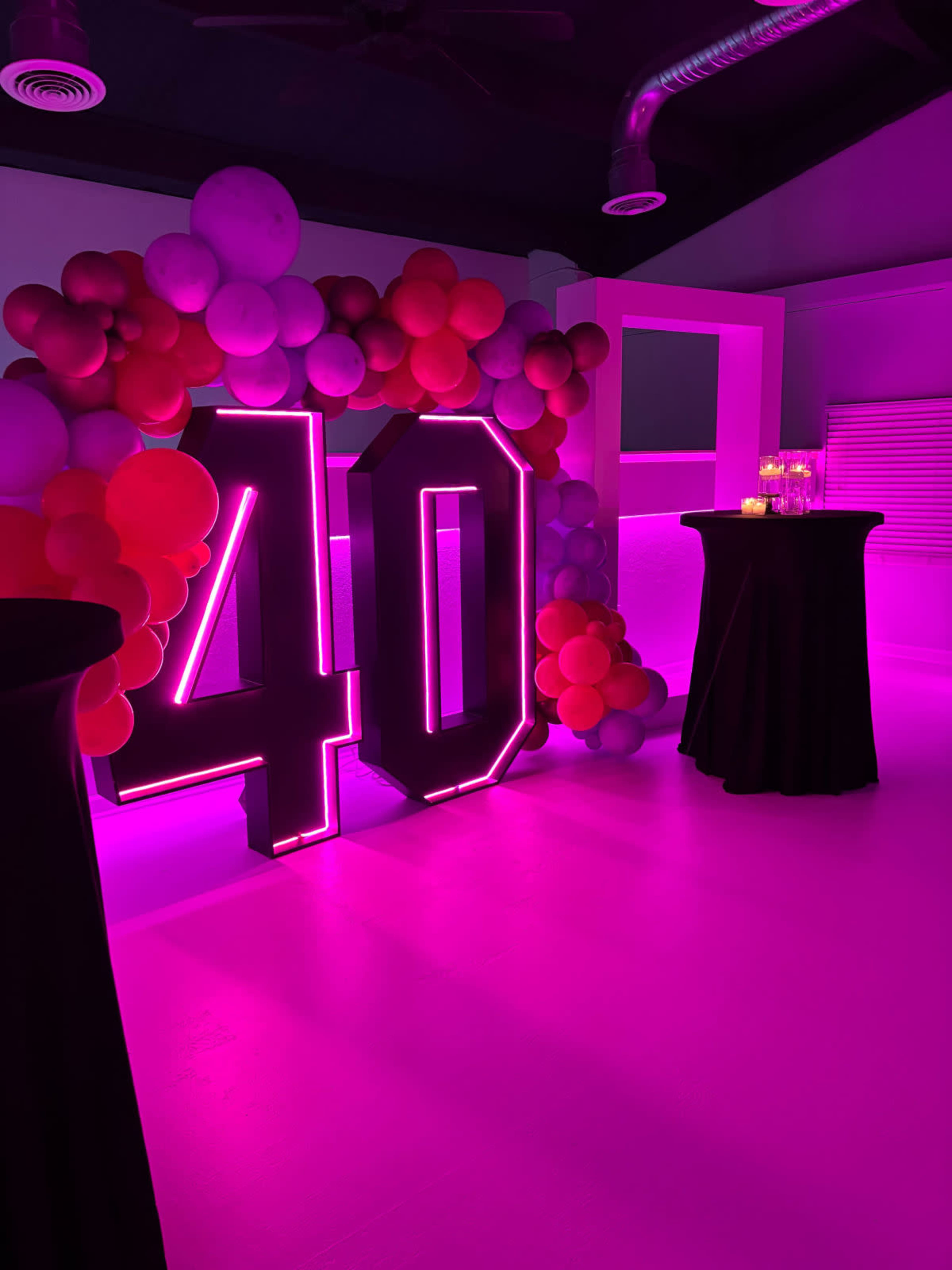 The image shows a decorated indoor space featuring a large illuminated "40" surrounded by colorful balloons, with black tables and soft lighting in shades of pink.