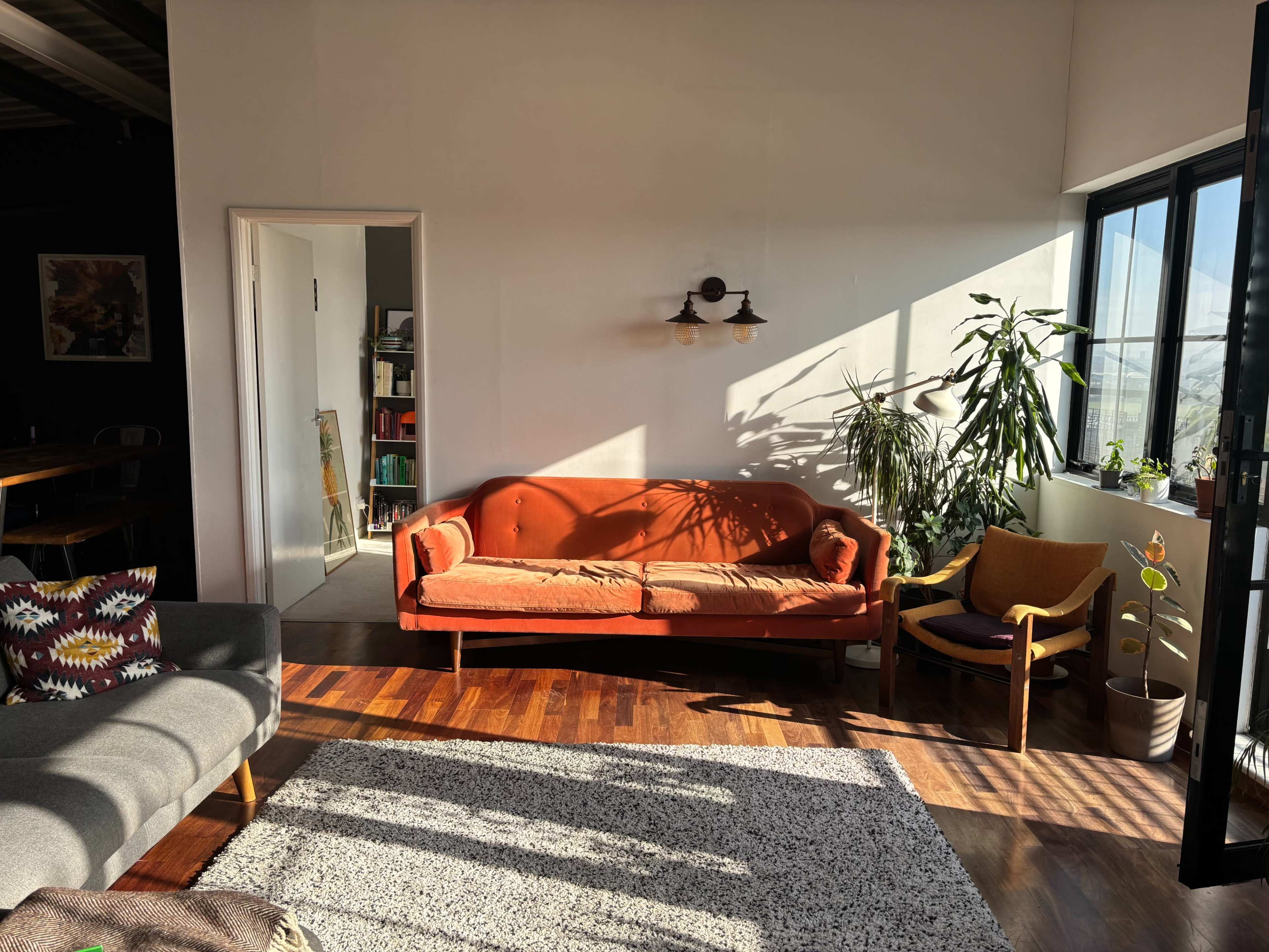 The scene shows a bright living room with an orange sofa, a wooden floor, and plants near large windows, with sunlight casting shadows across the space.