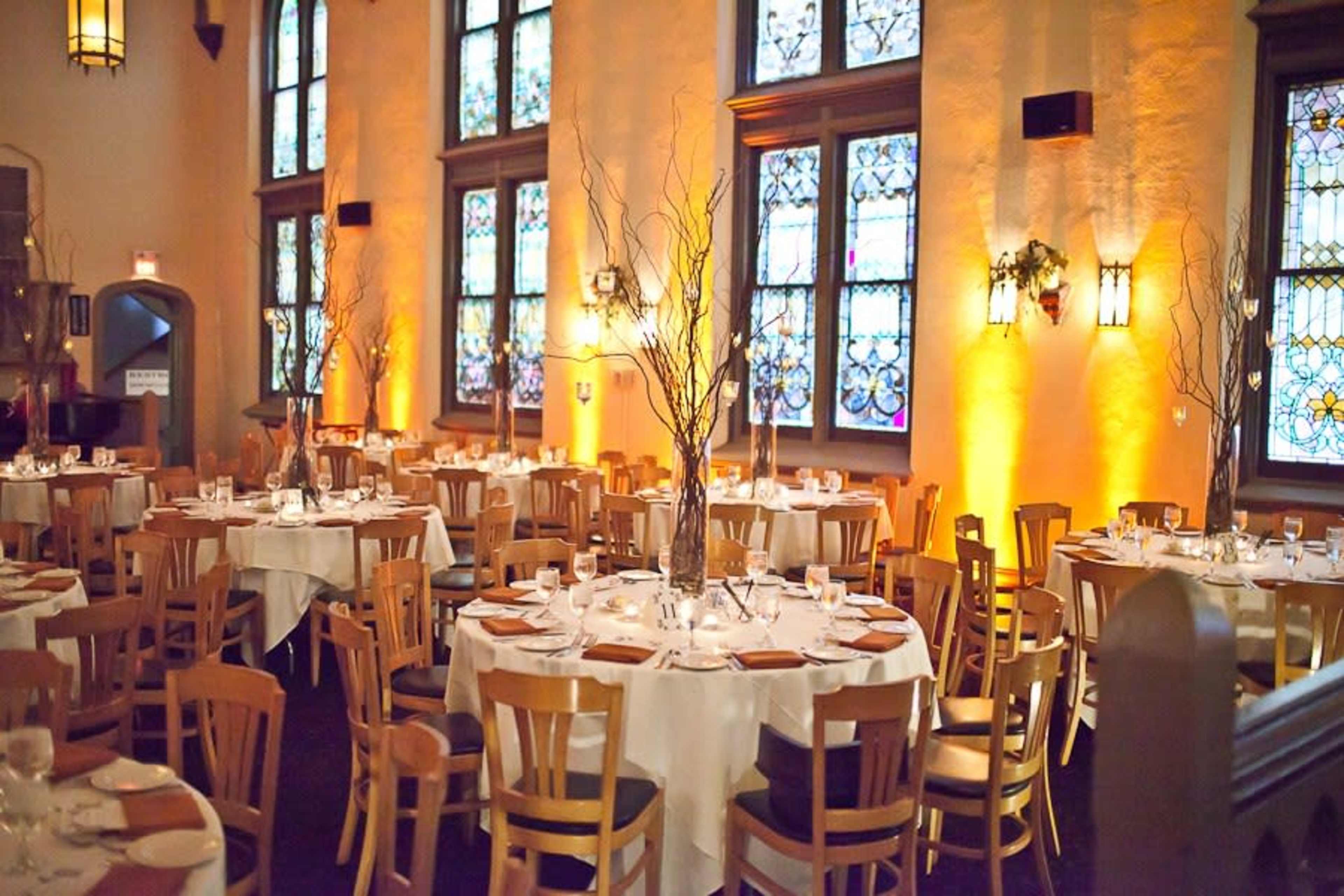 The image shows a dining hall with round tables set for a formal meal, surrounded by stained glass windows and warm lighting.