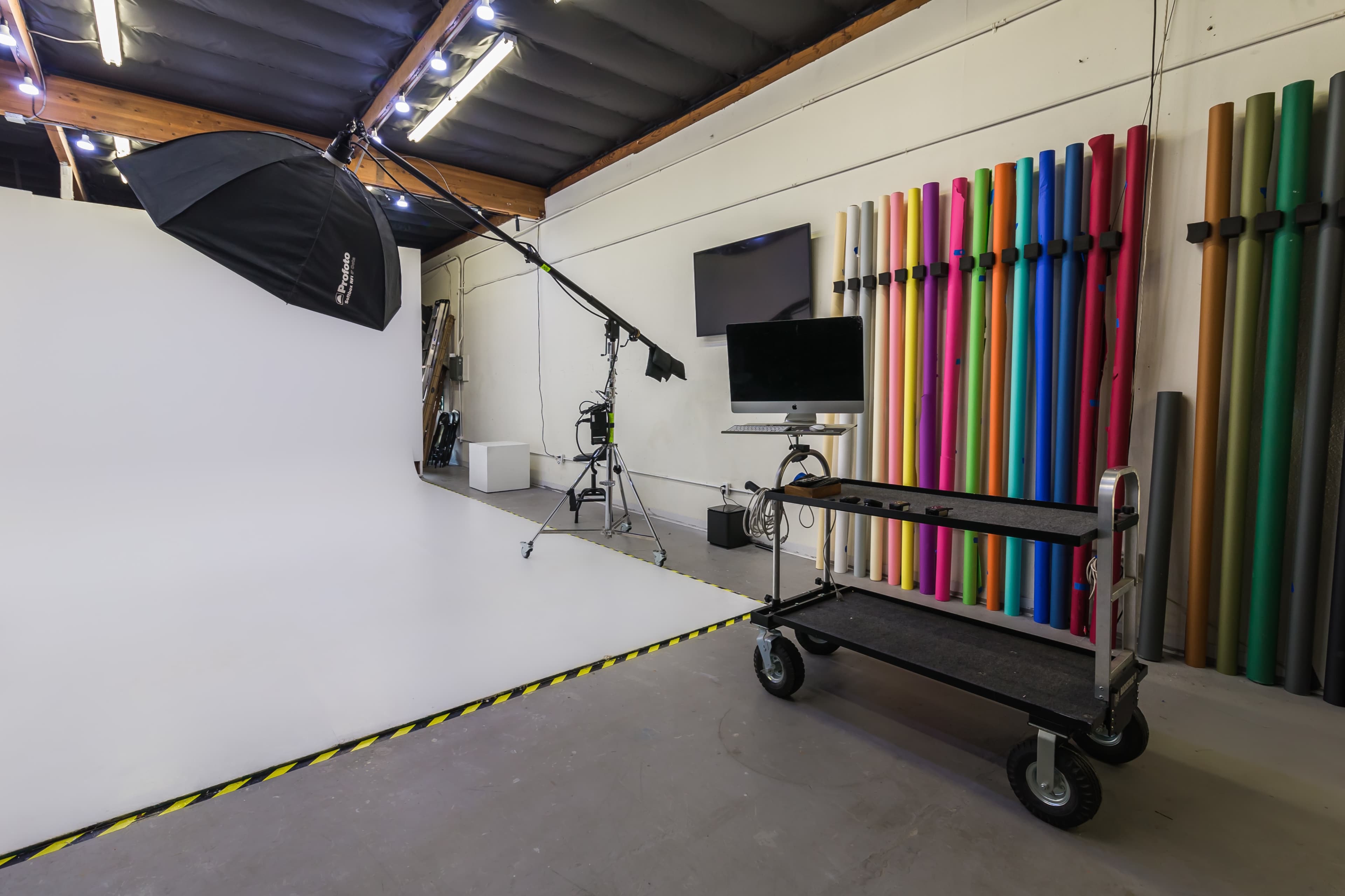 The image shows a photography studio with a white backdrop, lighting equipment, and a cart positioned near colorful rolls of seamless backdrops along one wall.