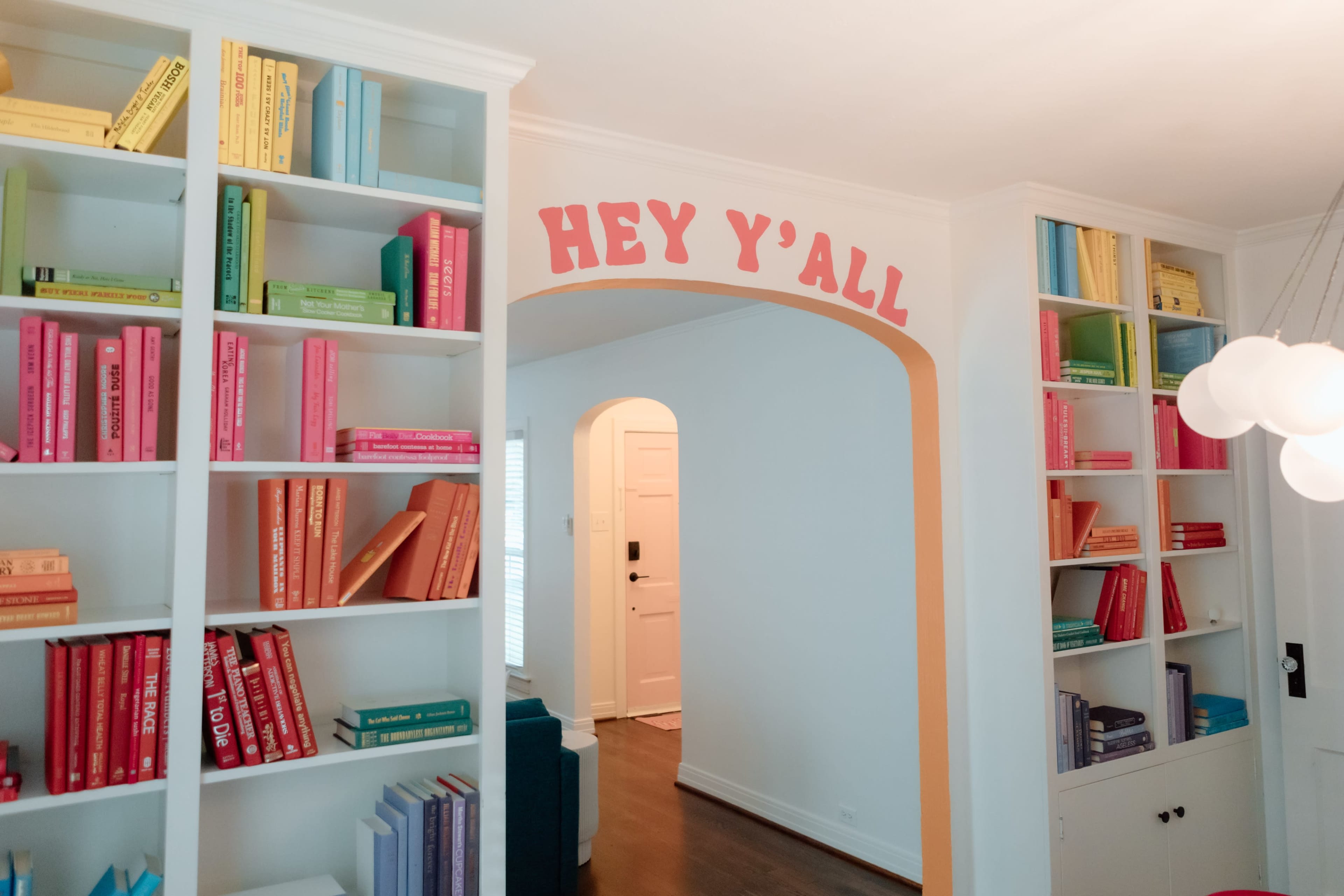 A bright room features two white bookshelves filled with colorful books and an archway with the text "HEY Y'ALL" painted above it.