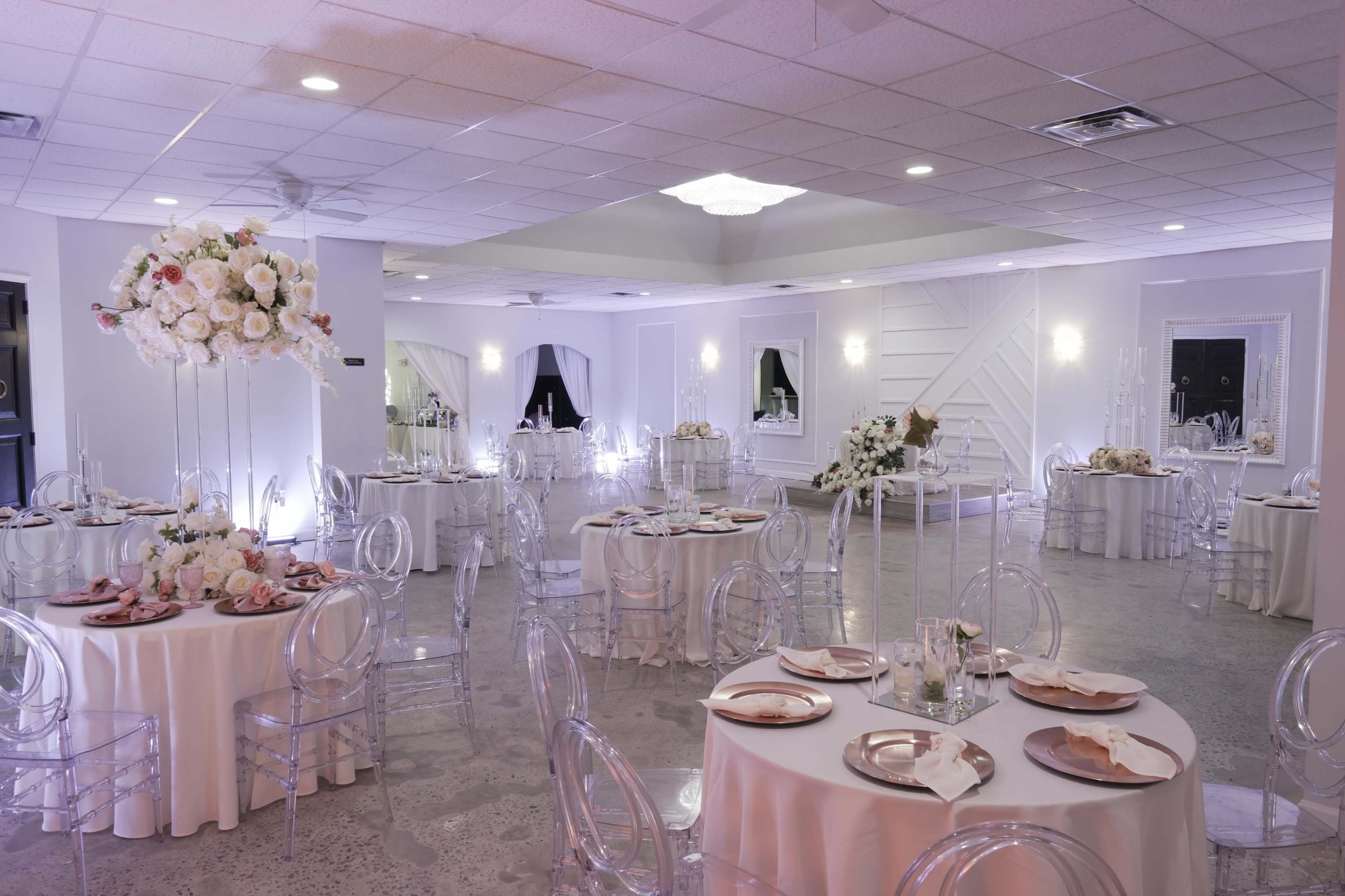 The image shows a spacious event hall set with round tables adorned with white tablecloths and elegant floral centerpieces, illuminated by soft overhead lighting.