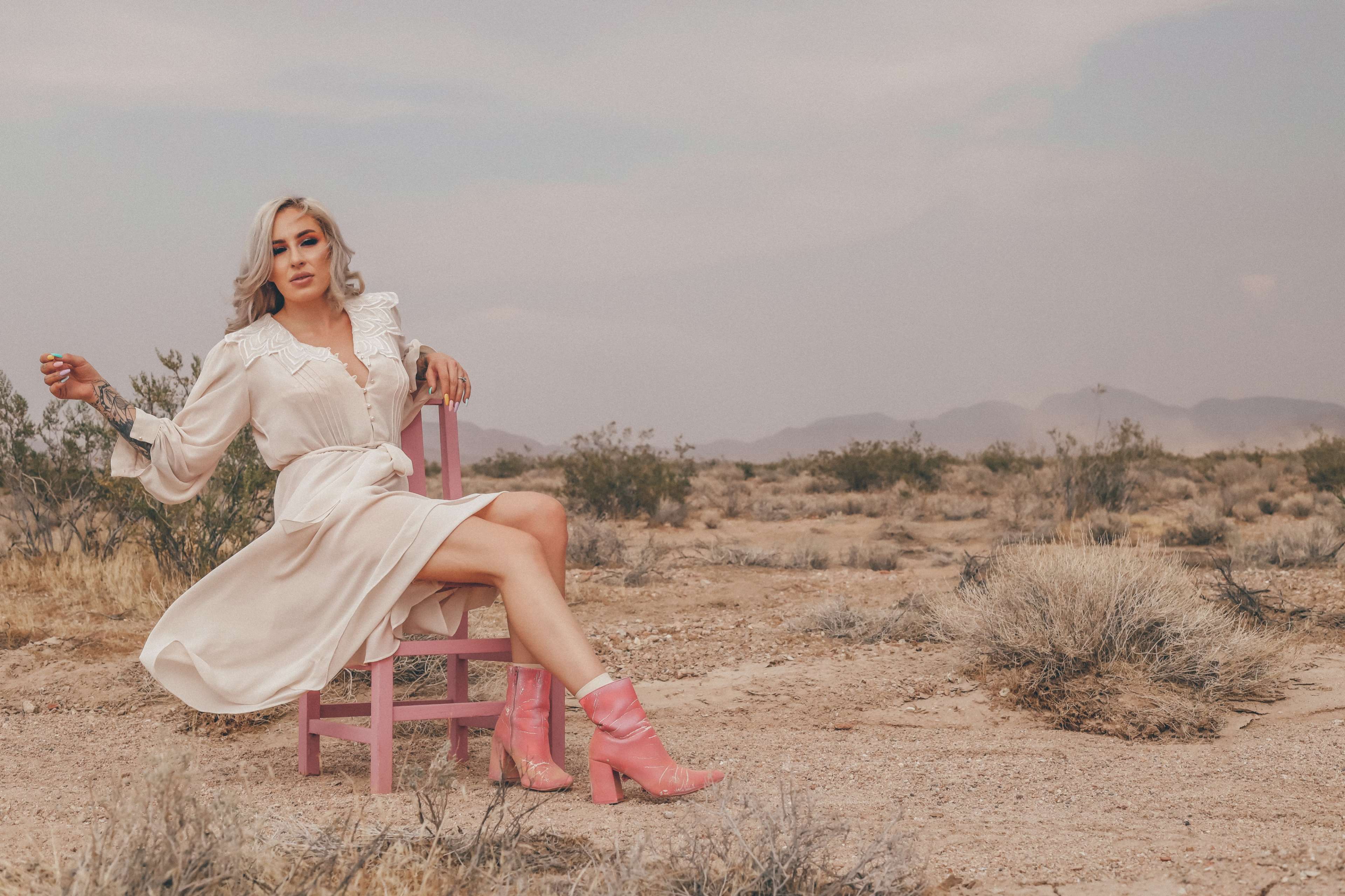 A woman in a white dress and pink boots sits on a pink chair in a desert landscape with mountains in the background.