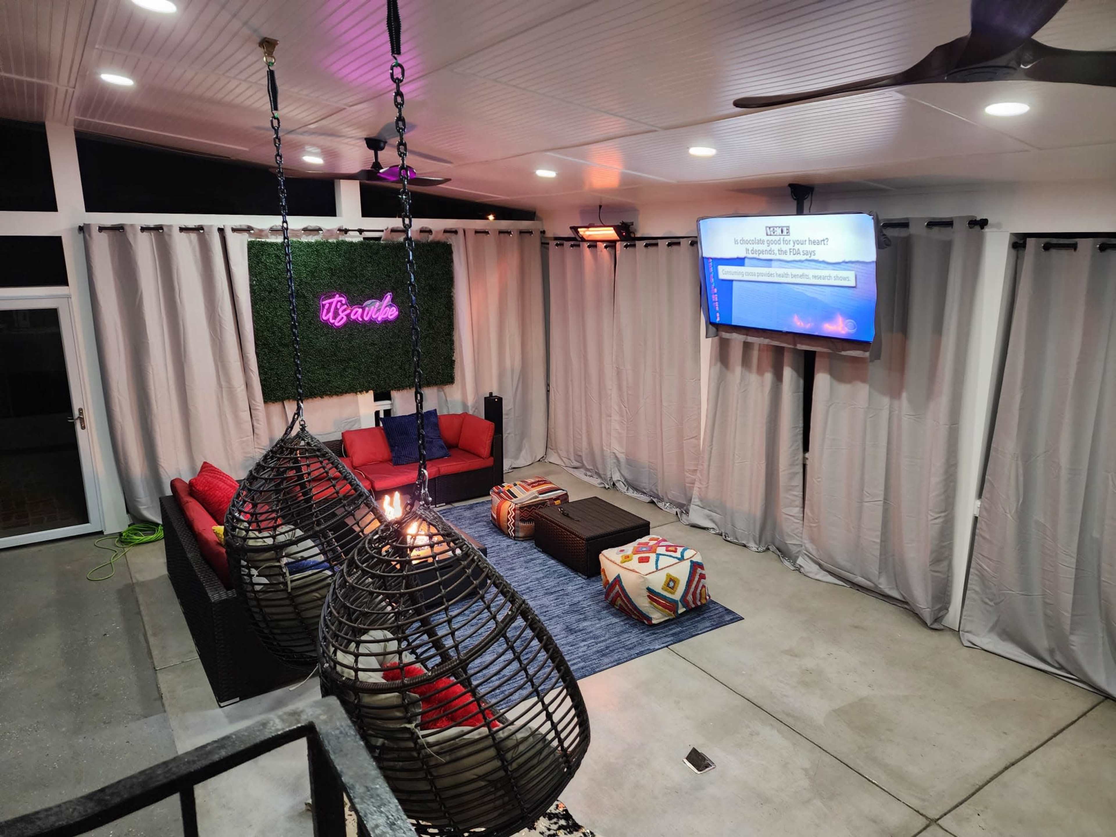 A modern outdoor lounge area with hanging chairs, a television on the wall, and a green plant backdrop.
