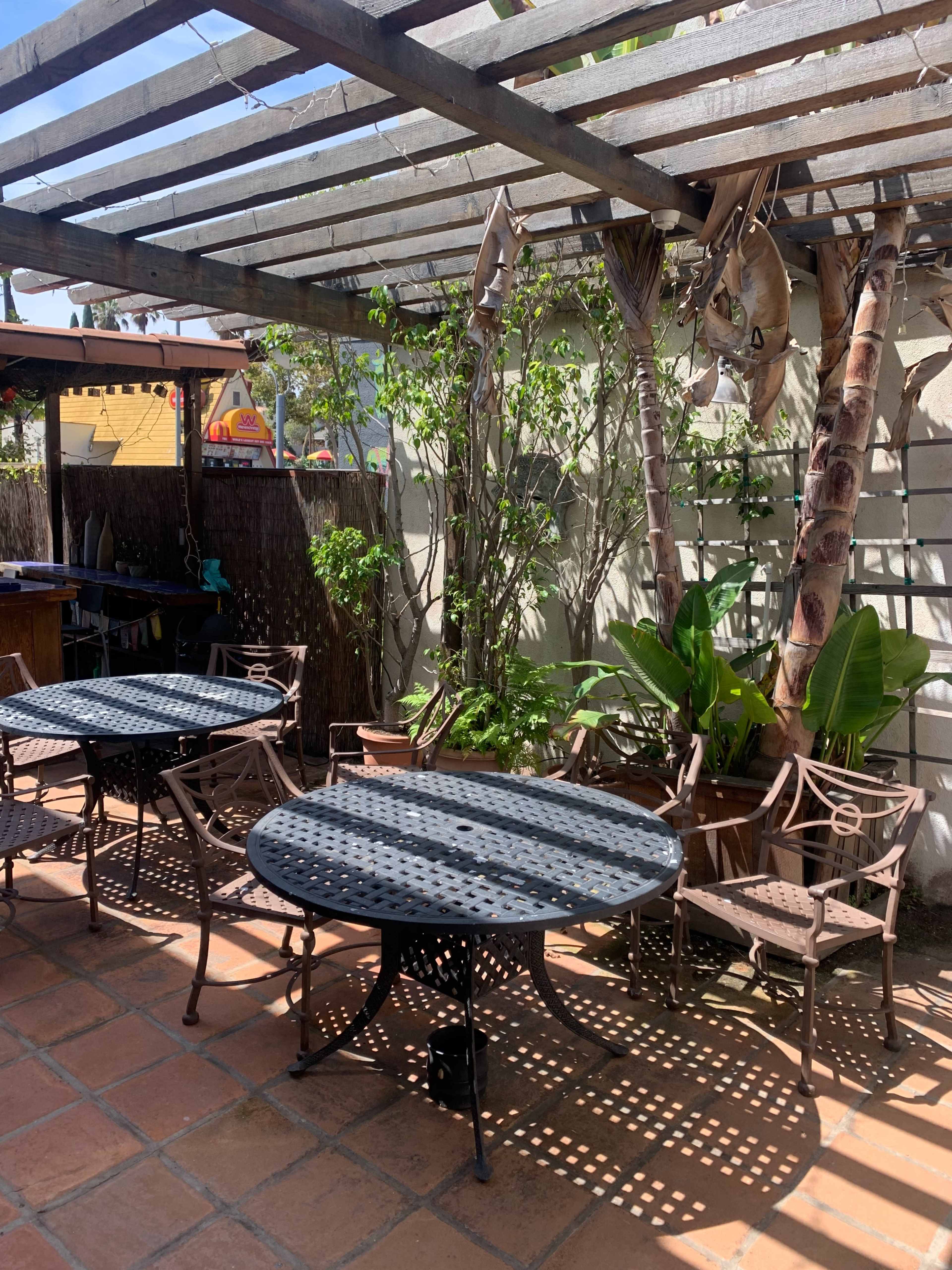 A shaded outdoor patio features multiple metal tables and chairs surrounded by potted plants and a pergola.