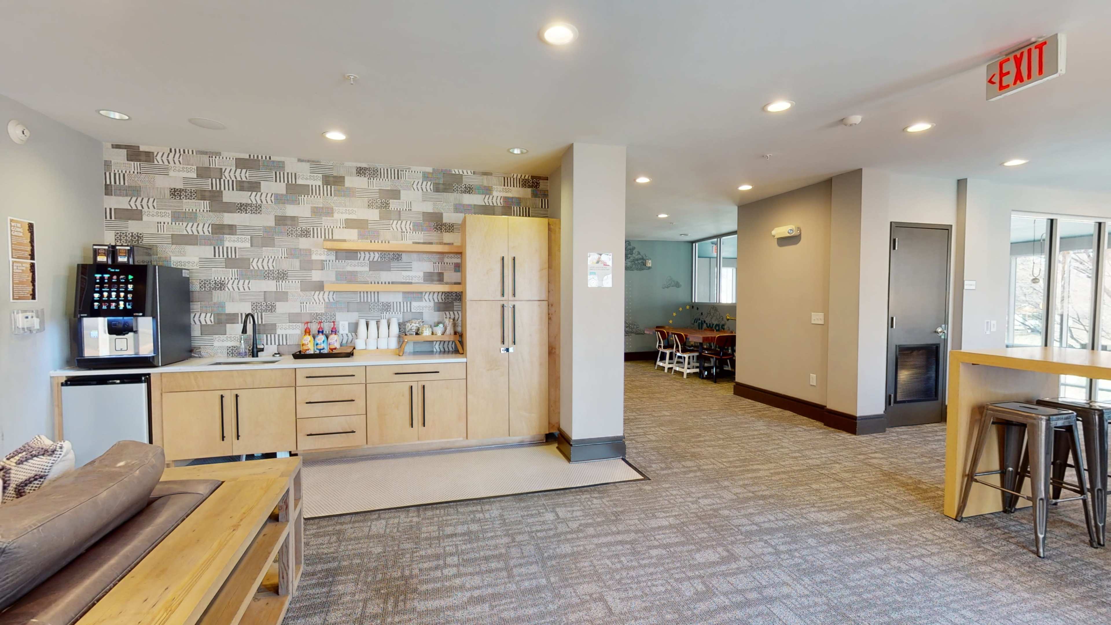 A modern common area with a kitchenette featuring wooden cabinetry, a coffee station, and seating arrangements.