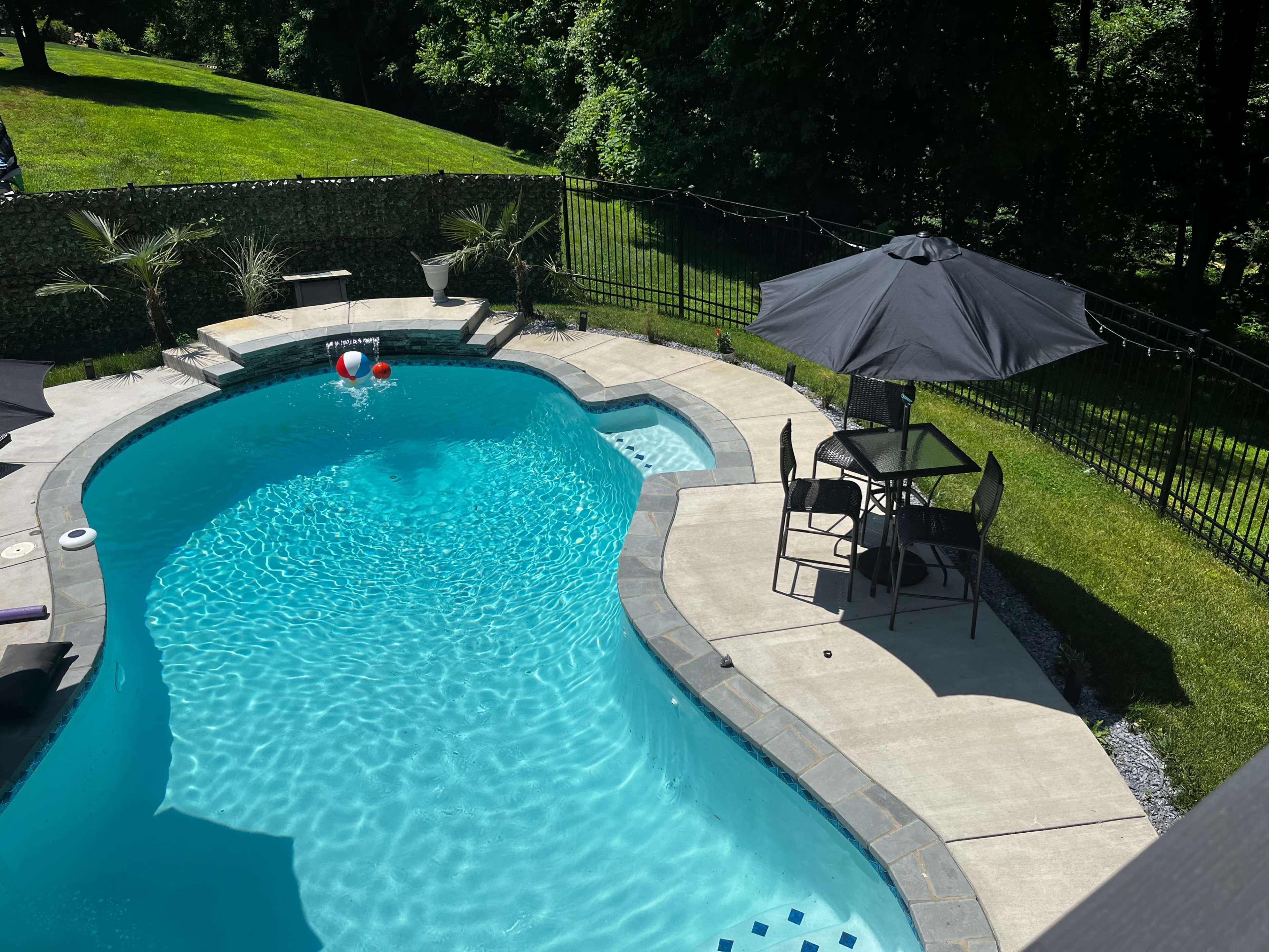 A backyard swimming pool surrounded by a concrete patio, lawn, and a table with chairs under a black umbrella.