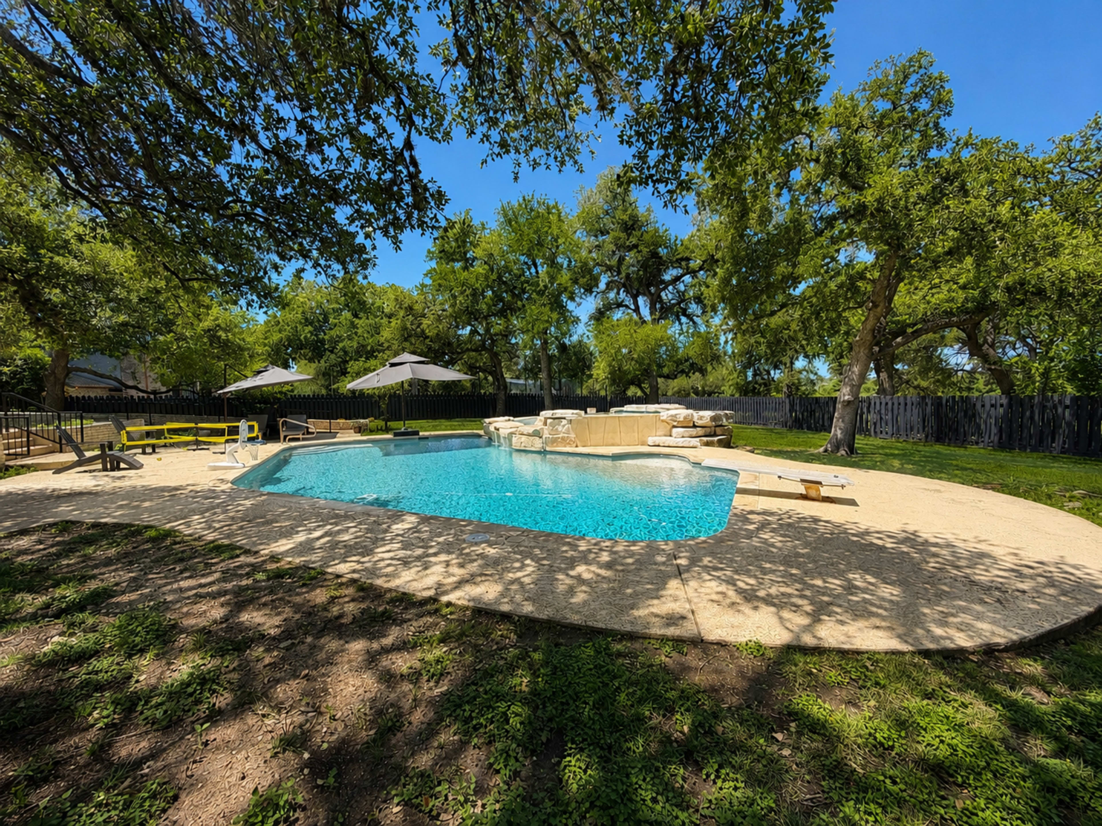 The image shows a swimming pool surrounded by a patio and shaded by trees, with lounge chairs and umbrellas nearby.