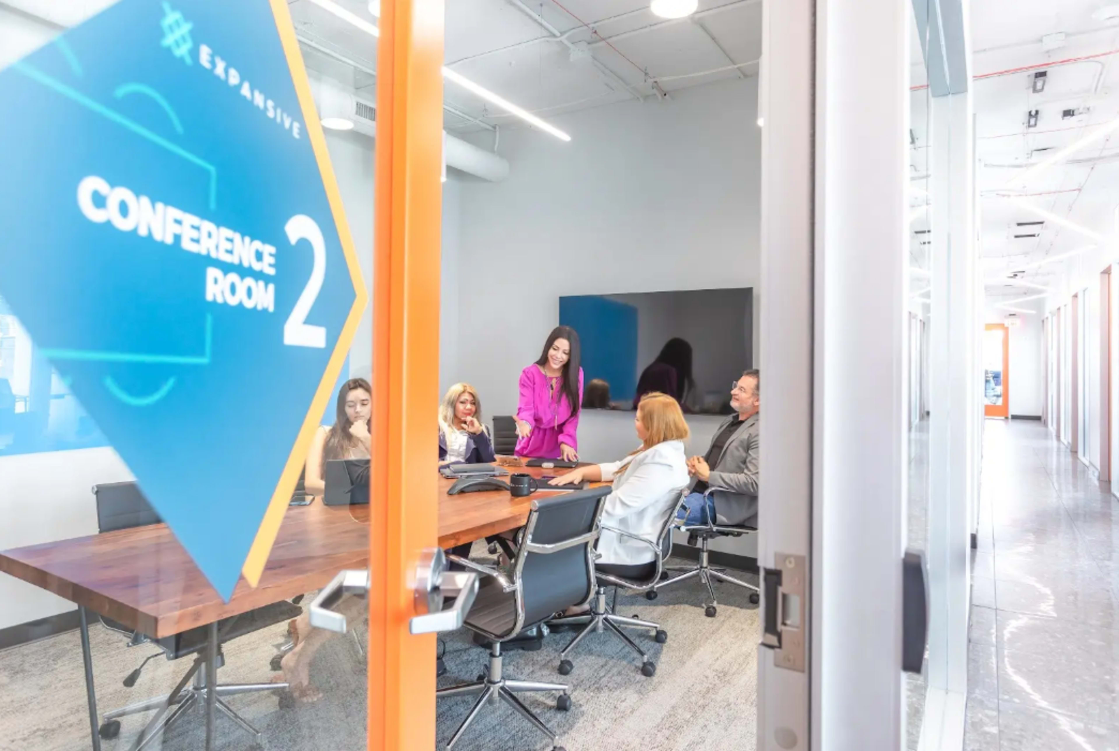 A group of five people is seated around a conference table in a modern conference room, while a woman stands and engages with them.