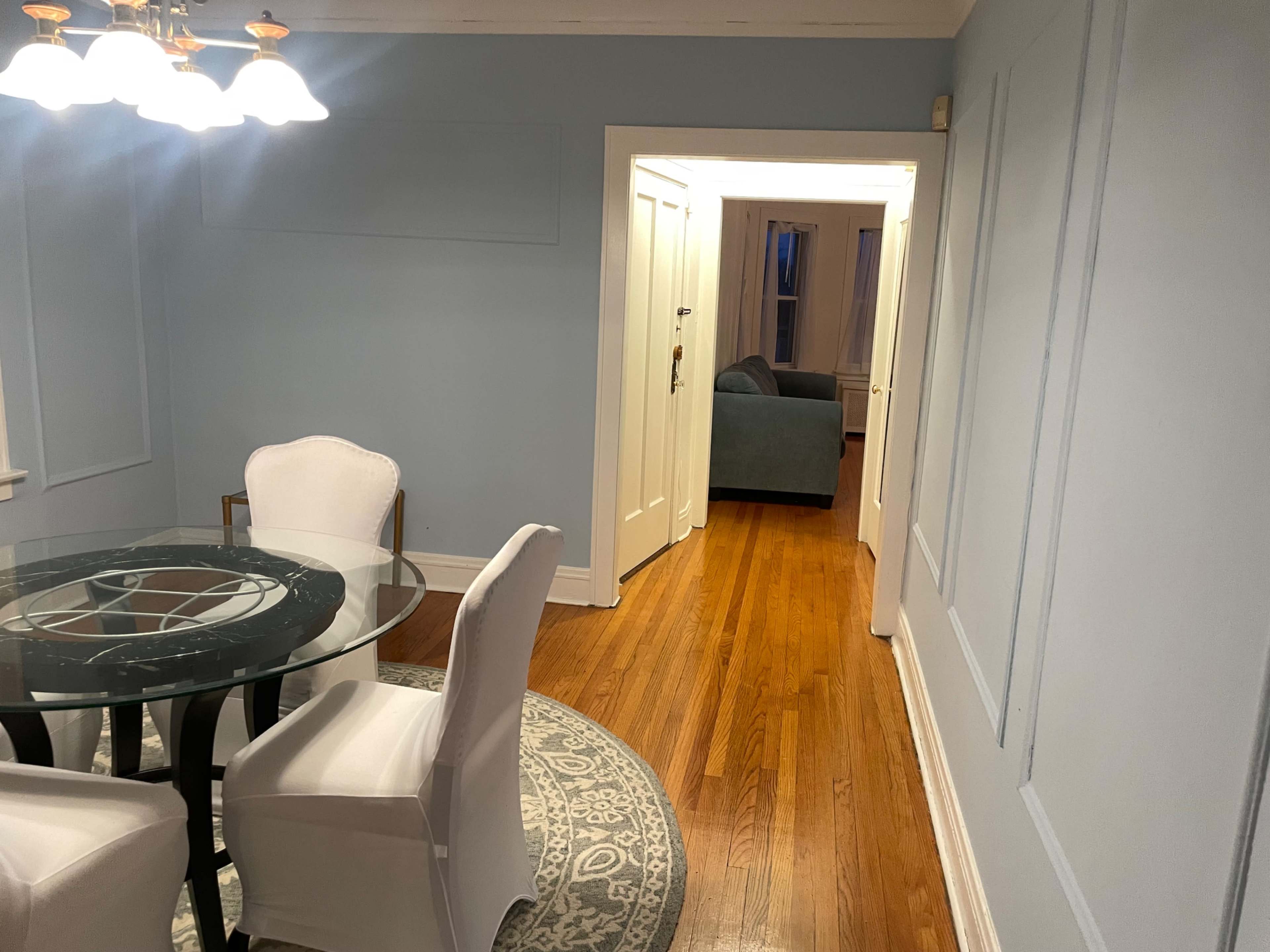 The image shows a dining area with a round glass table and white chairs, adjacent to a hallway leading to another room.