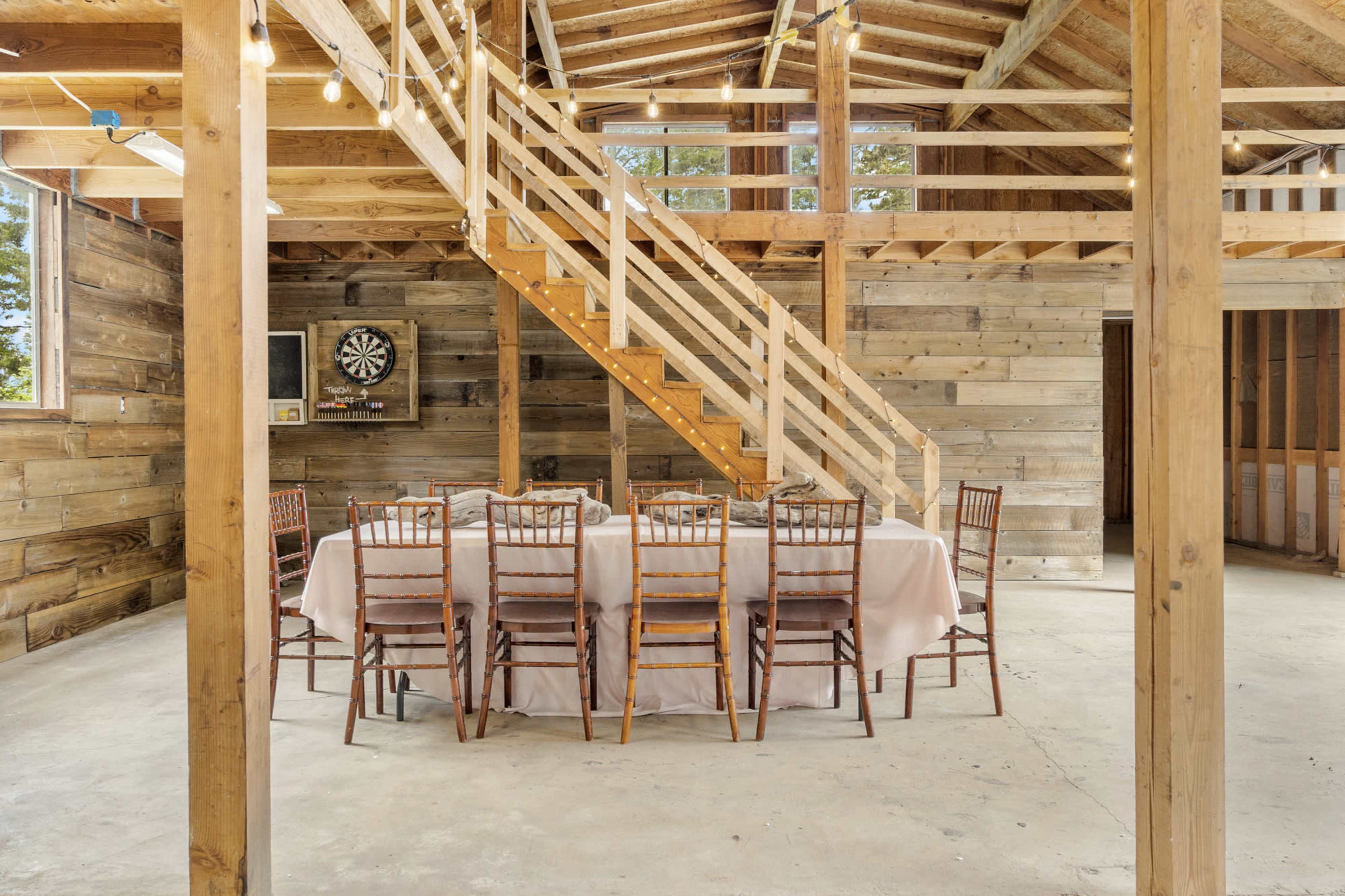 The image shows a rustic interior of a wooden barn featuring a large round table set for dining, surrounded by chairs, with a staircase leading to an upper level.