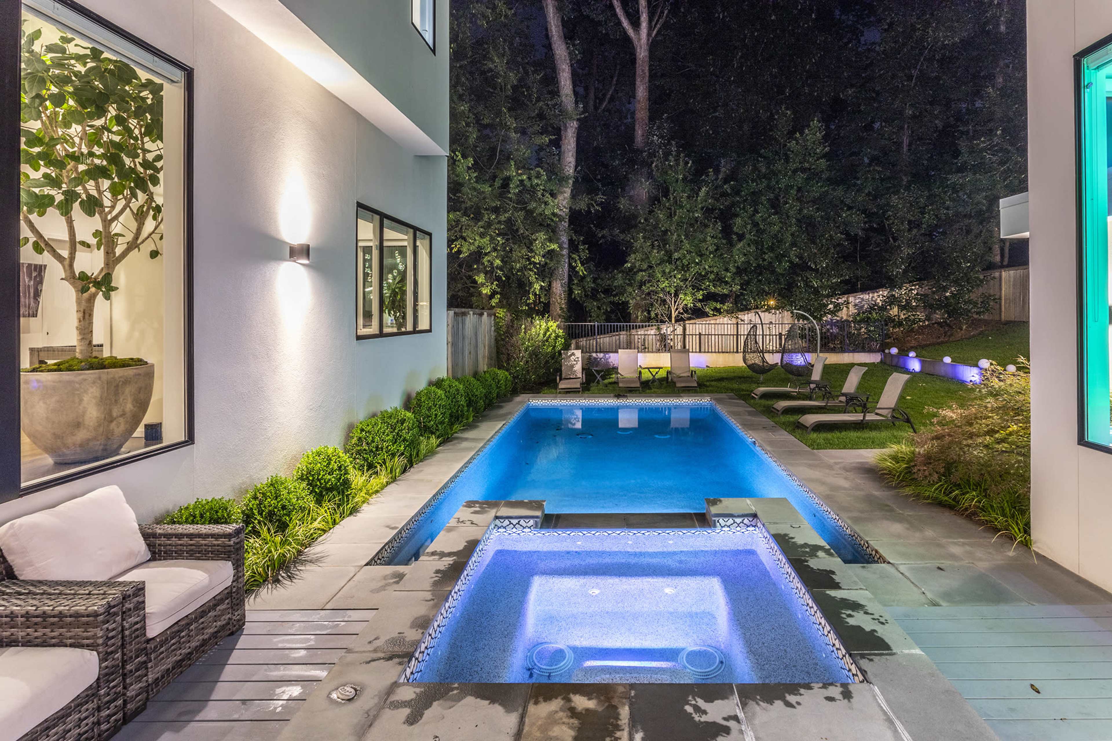 A modern backyard with a rectangular pool, a hot tub, and several lounge chairs surrounded by greenery at night.