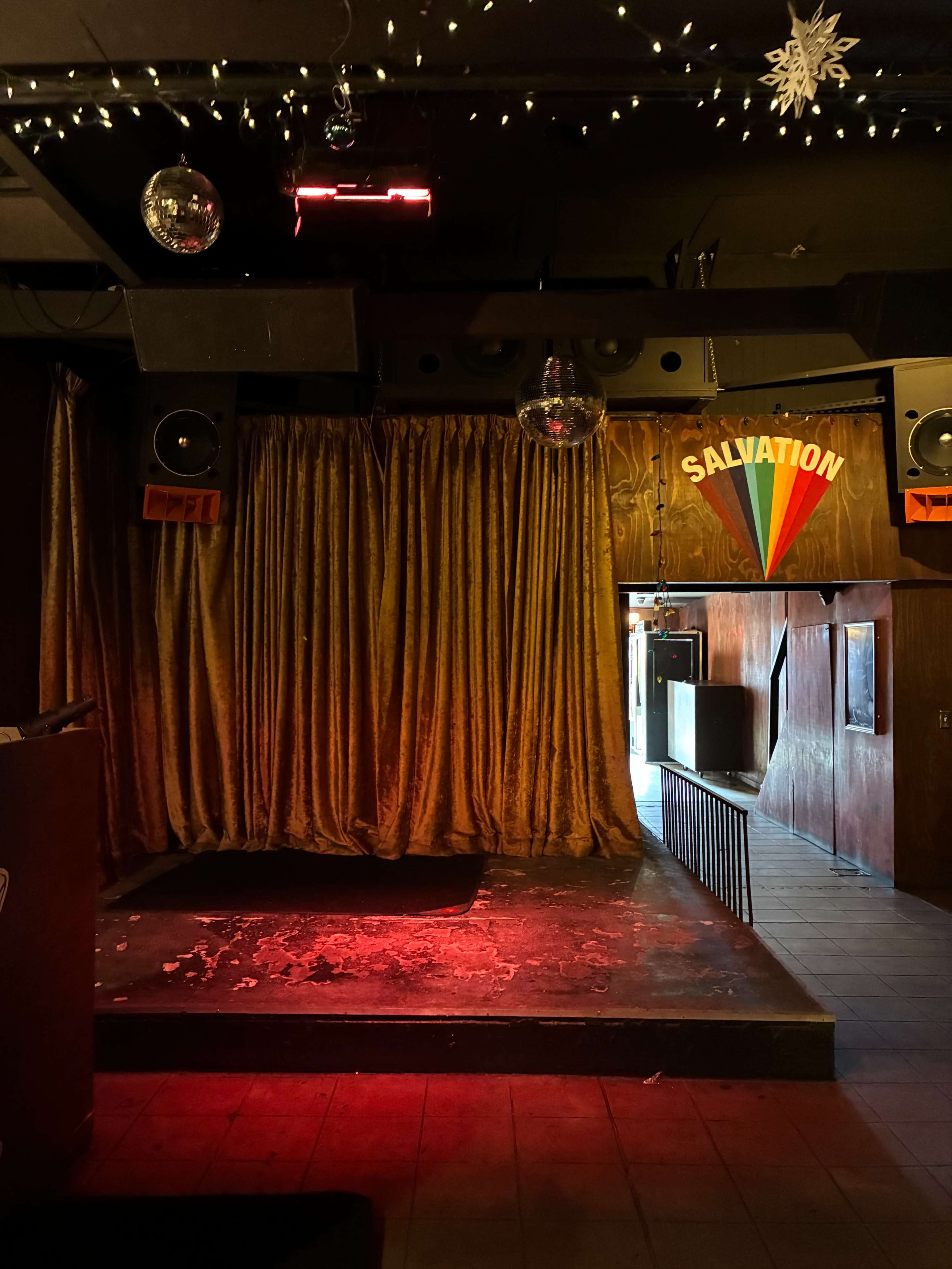 The image shows a dimly lit stage with a red curtain and a sign that reads "SALVATION," decorated with disco balls and overhead string lights.