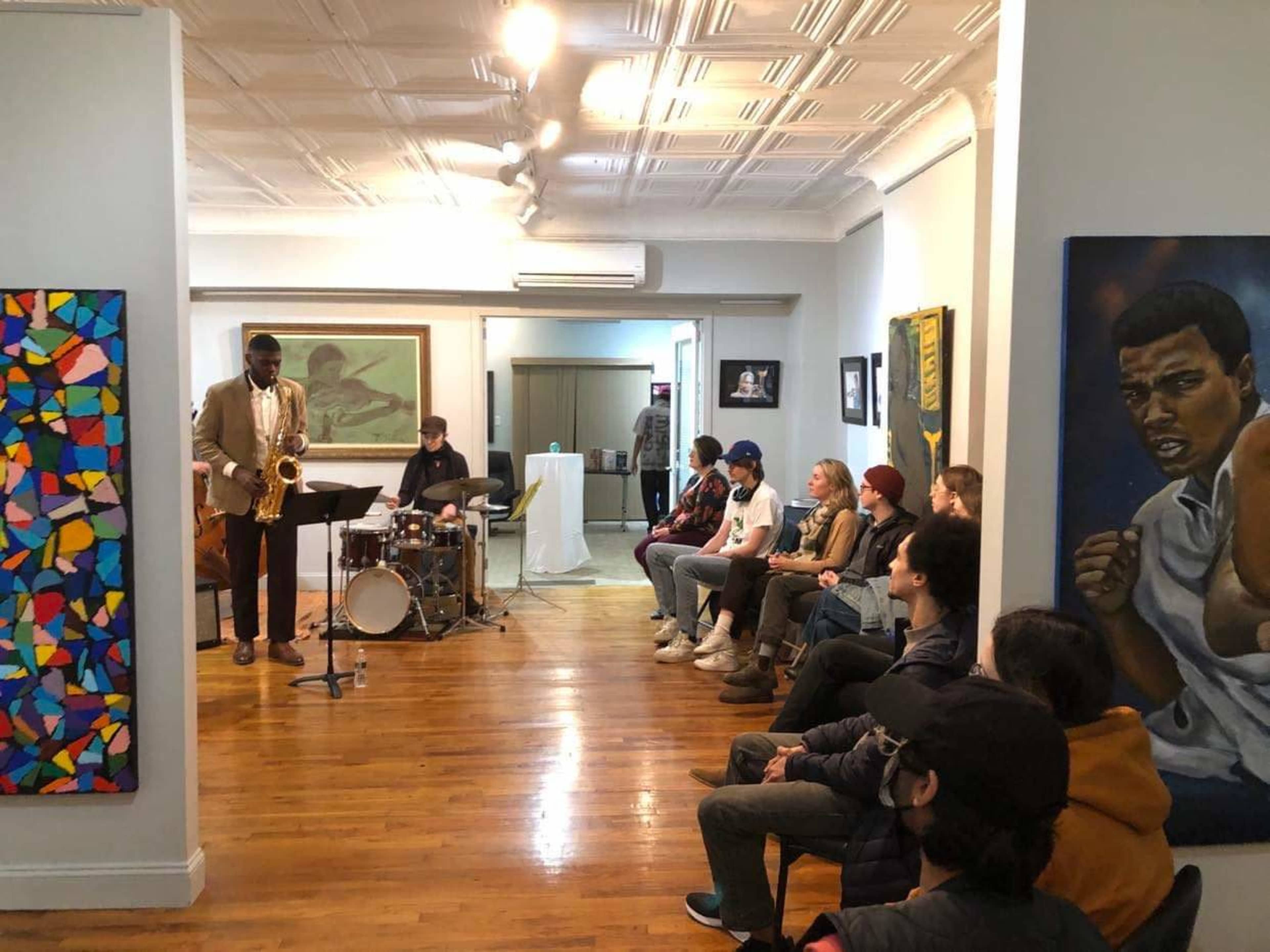 A saxophonist plays in an art gallery while a drummer accompanies him, as an audience of various individuals observes from the seating area.