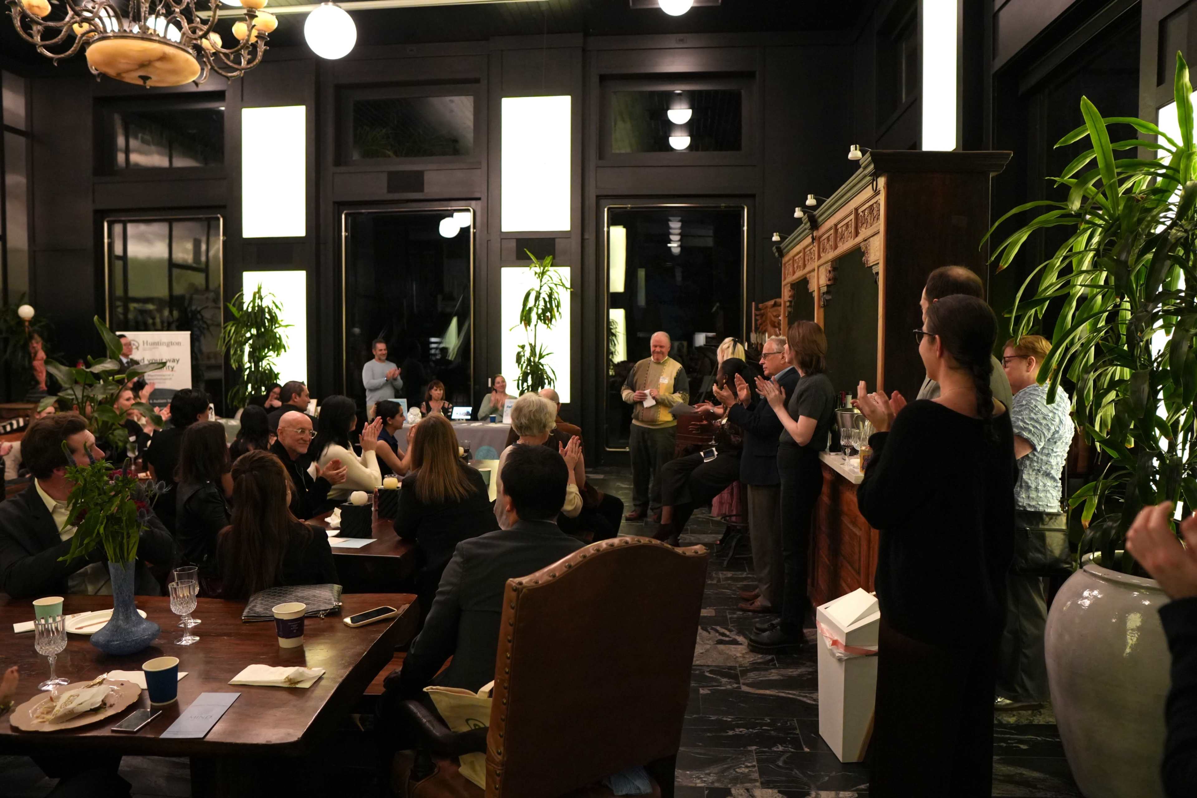 A group of people is gathered in a well-lit room with large windows, seated around tables while some stand and engage in conversation.